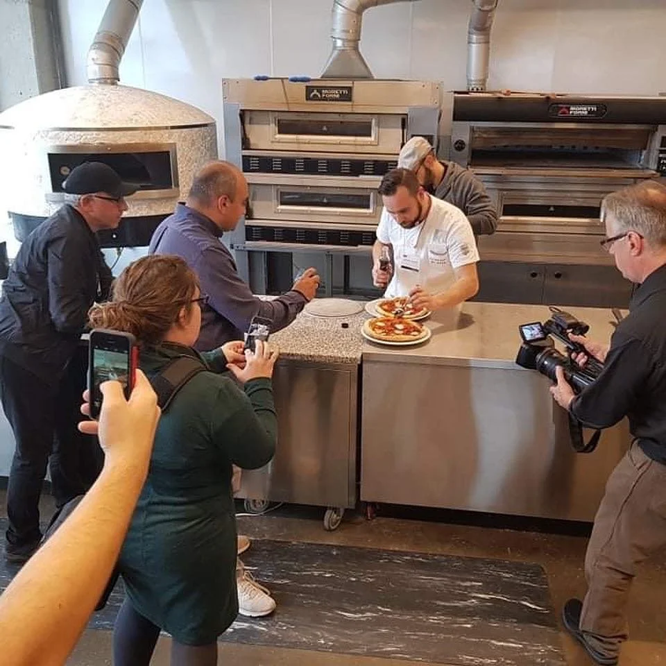 Group of people filming a chef preparing pizzas in a commercial kitchen with large ovens in the background.