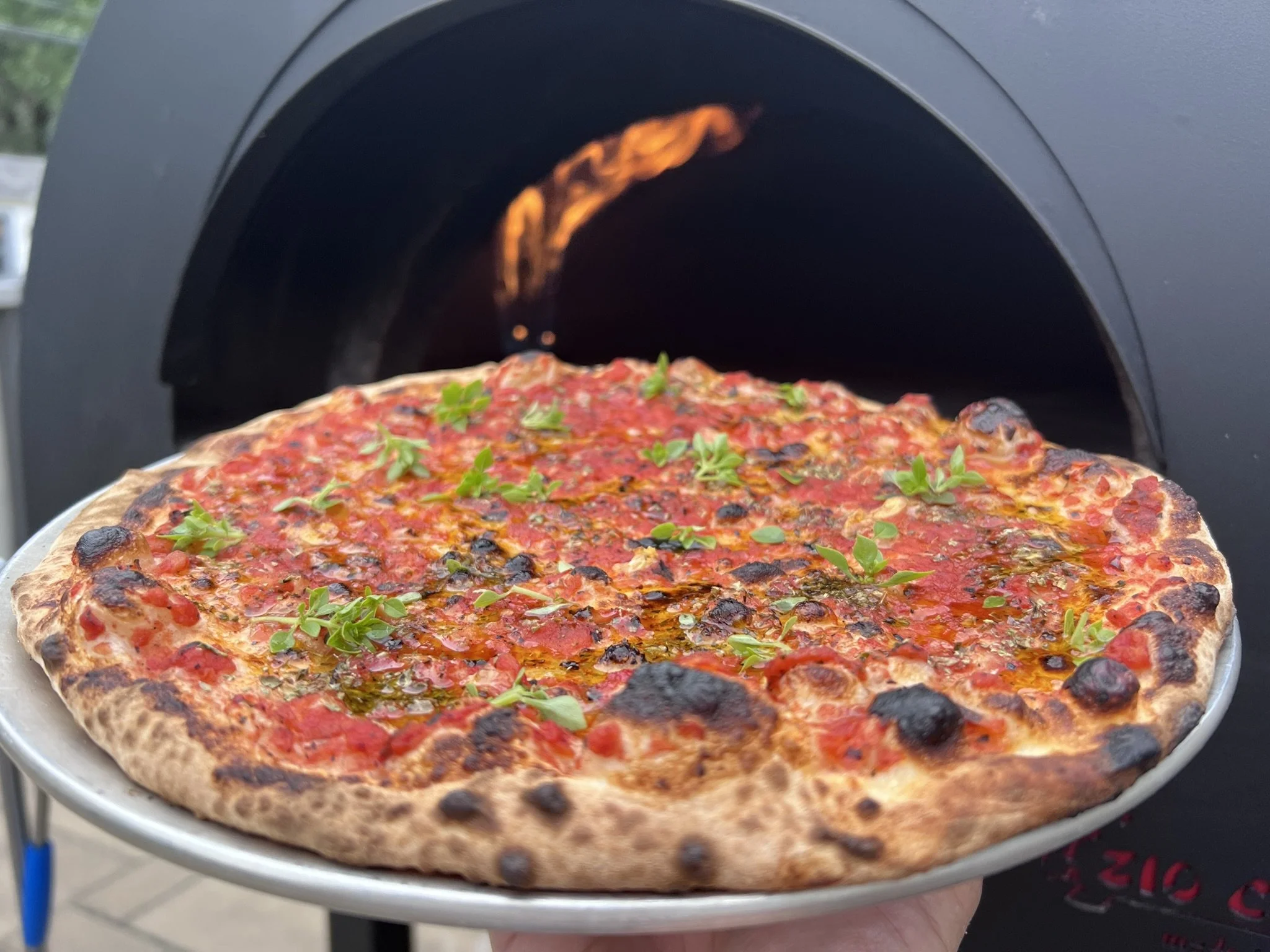 A freshly baked Marinara pizza with tomatoes, herbs, and garlic facing a black pizza oven.