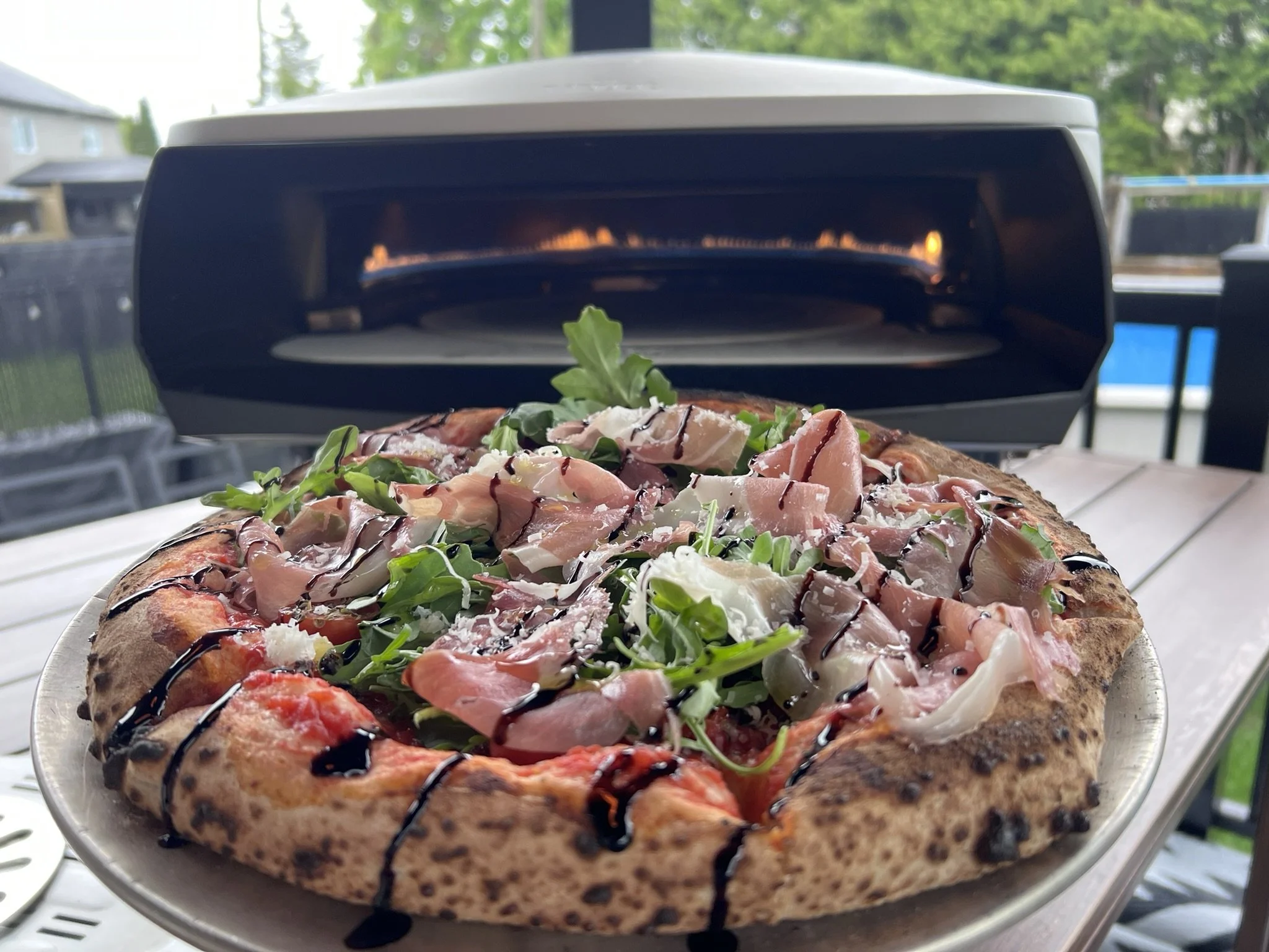 Pizza with arugula, prosciutto, cheese, and balsamic glaze on a wooden table outdoors.