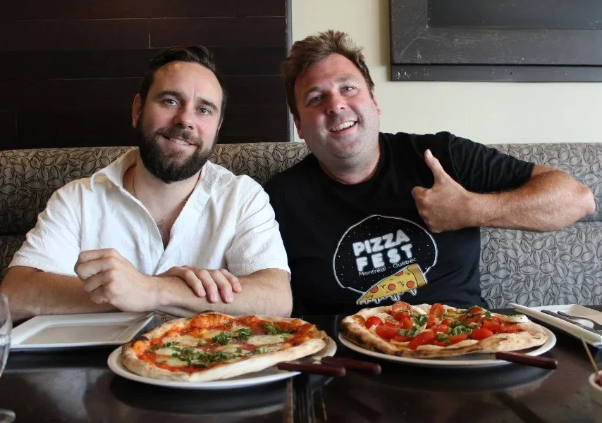Two men sitting at a restaurant table with two pizzas in front of them, smiling at the camera, one giving a thumbs-up.