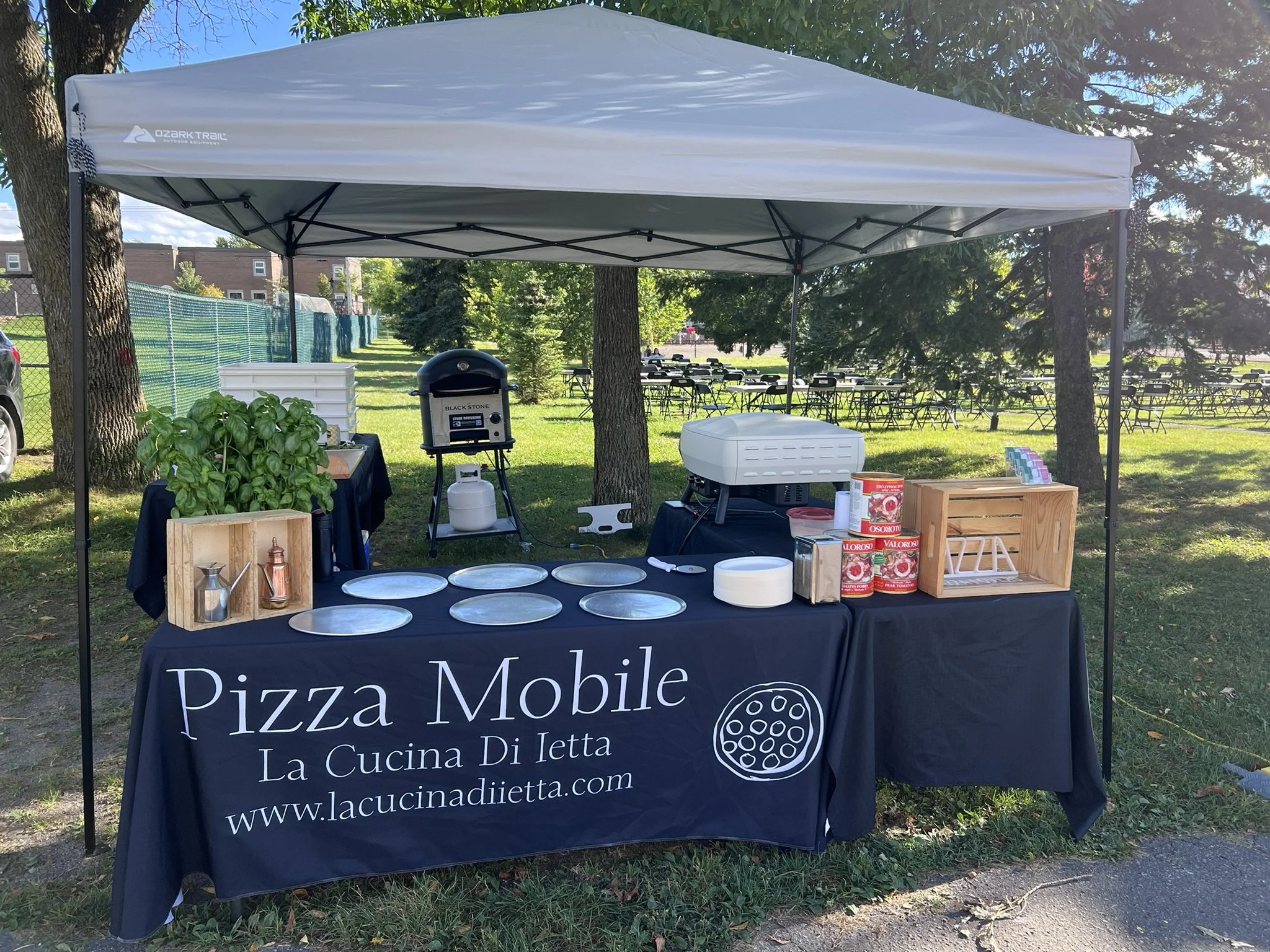 A pizza food stand under a canopy, featuring pizza-making equipment, plates, and ingredients, with a sign advertising "Pizza Mobile La Cucina Di Ietta" at an outdoor event in a park.