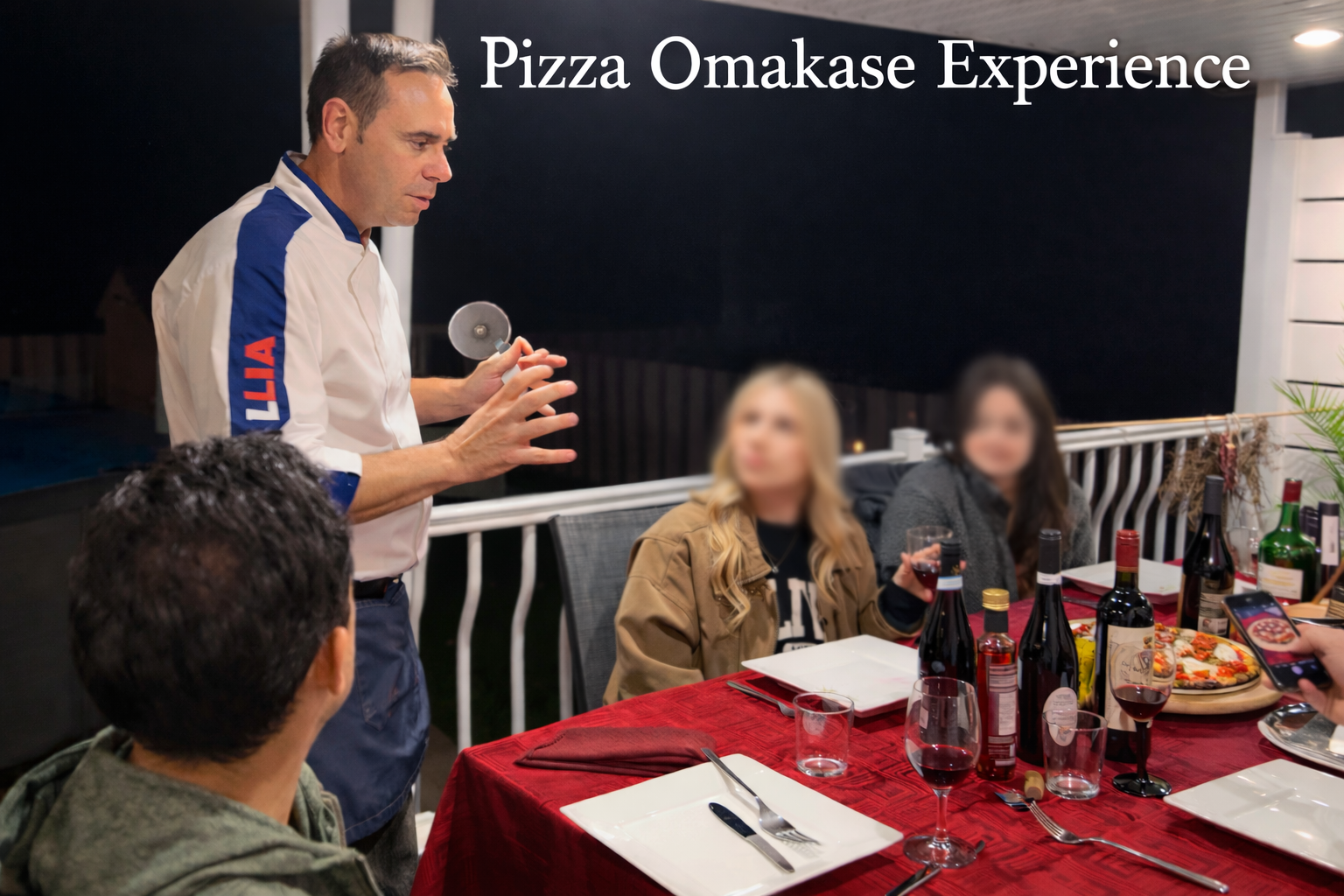 A man in a white and blue shirt demonstrating pizza making on a balcony at night for a group of people with wine bottles, glasses, and a pizza on the table.