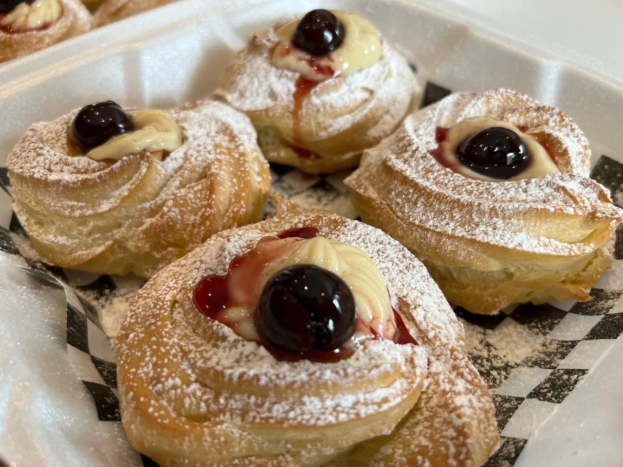 Four Zeppole di San Giuseppe topped with powdered sugar, custard cream, Amarena cherries, and syrup, arranged in a white basket lined with checkerboard paper.