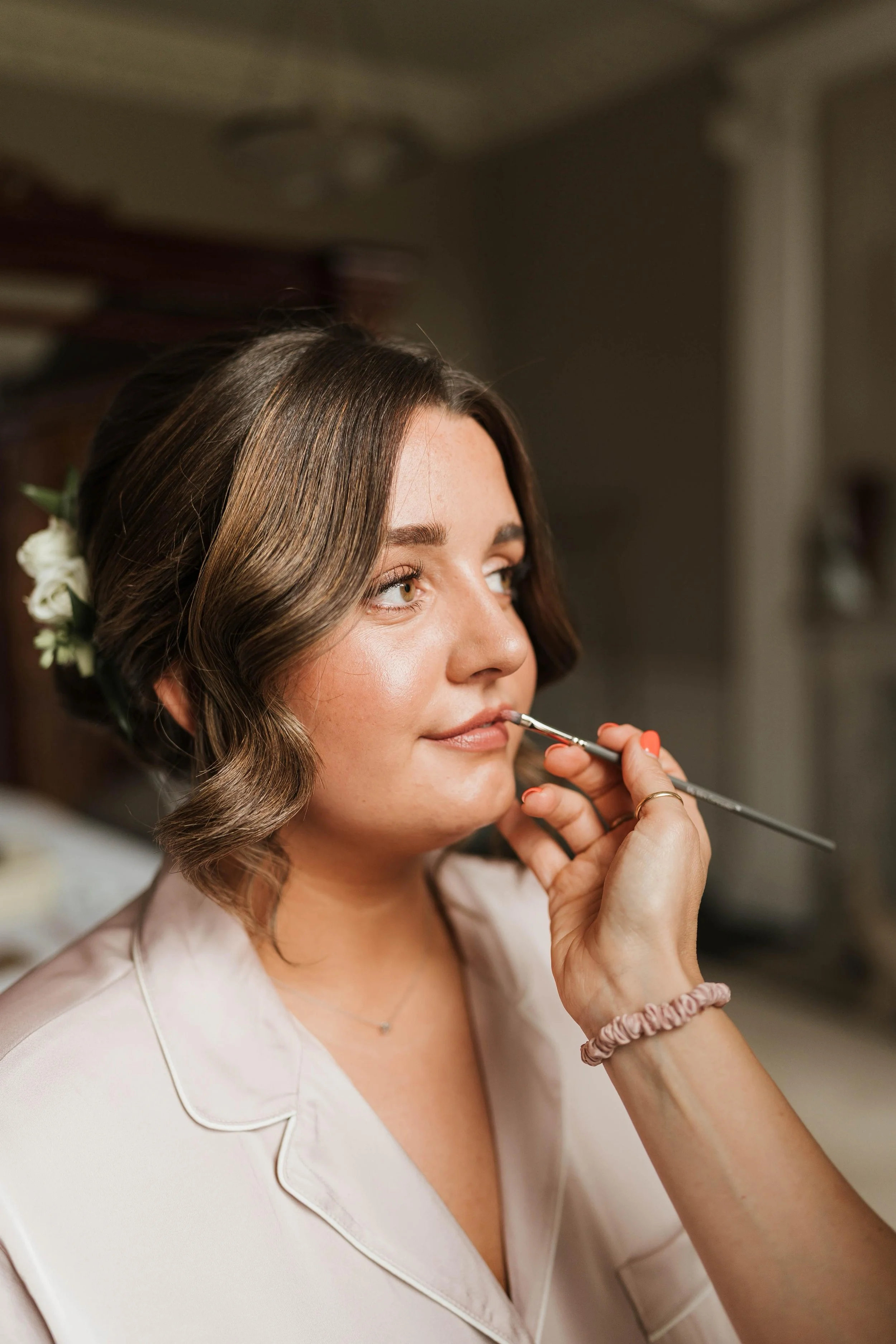 A woman with short, wavy brown hair and floral hair accessories is having her makeup done, with a makeup brush near her lips.