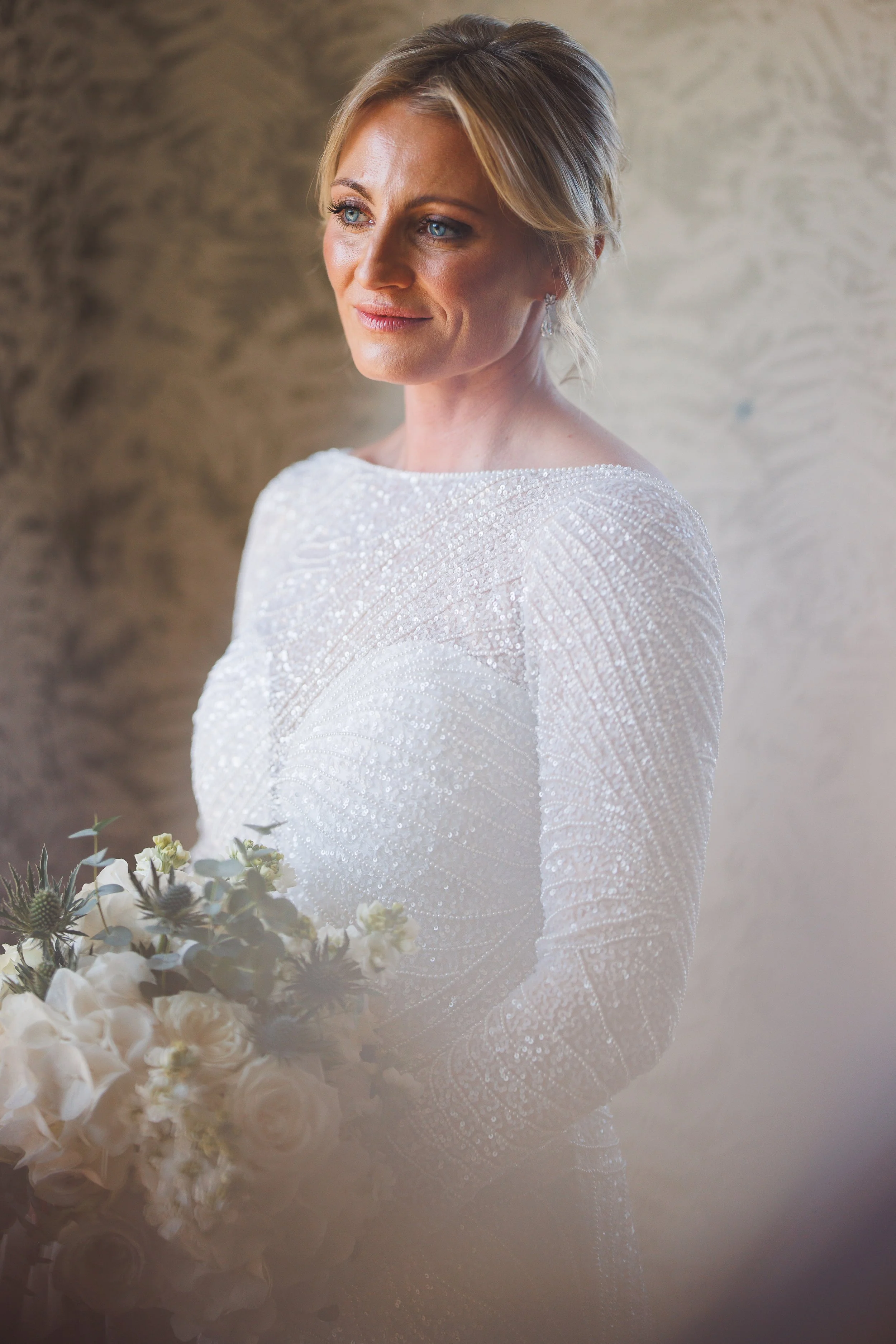A bride with blonde hair and blue eyes smiling slightly, wearing a long-sleeved, beaded white wedding dress, holding a bouquet of white flowers, standing in front of a textured beige wall.