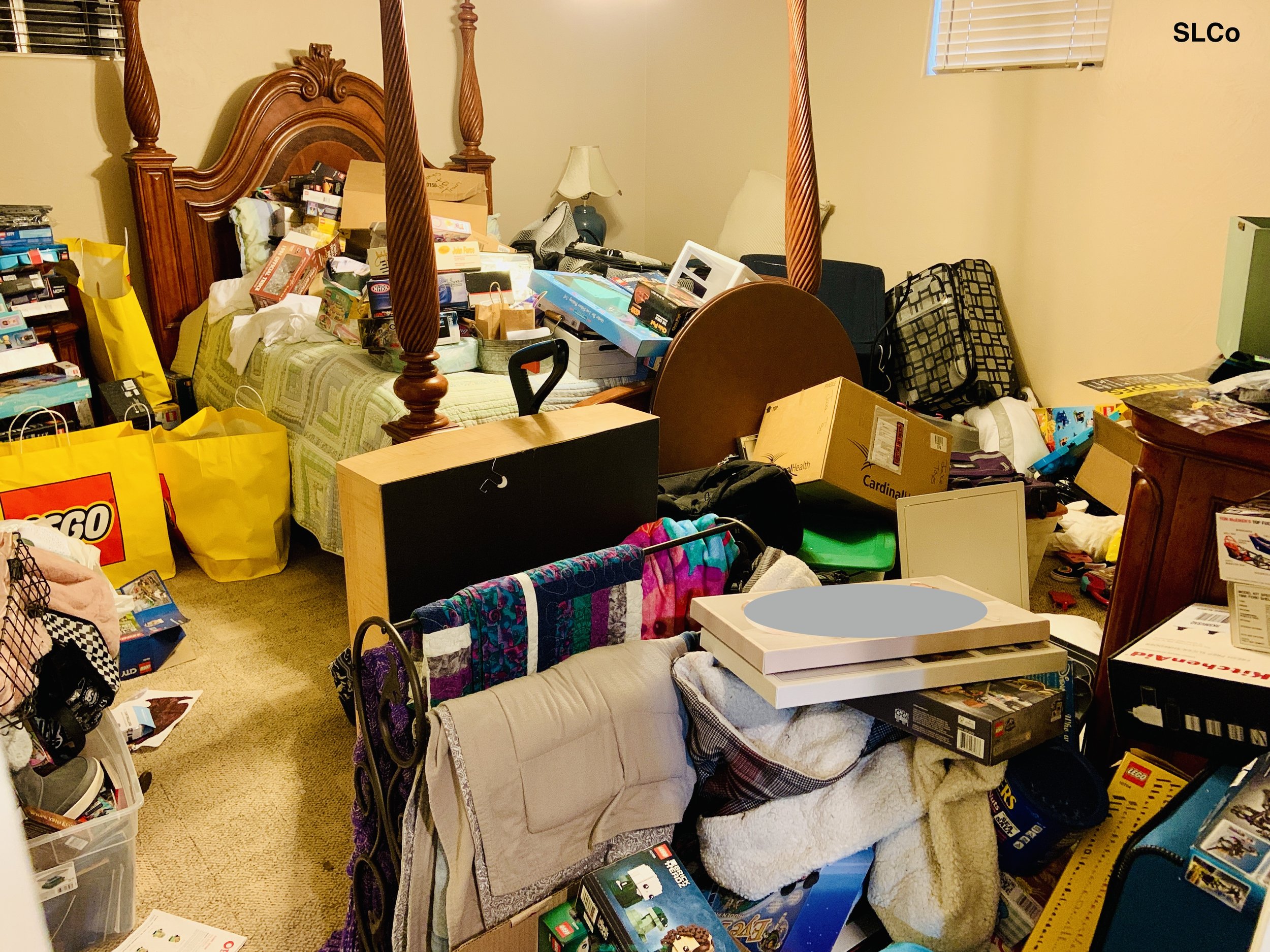 Before photo of bedroom with large bed with wooden posts, boxes and items on bed, floor covered with items and boxes