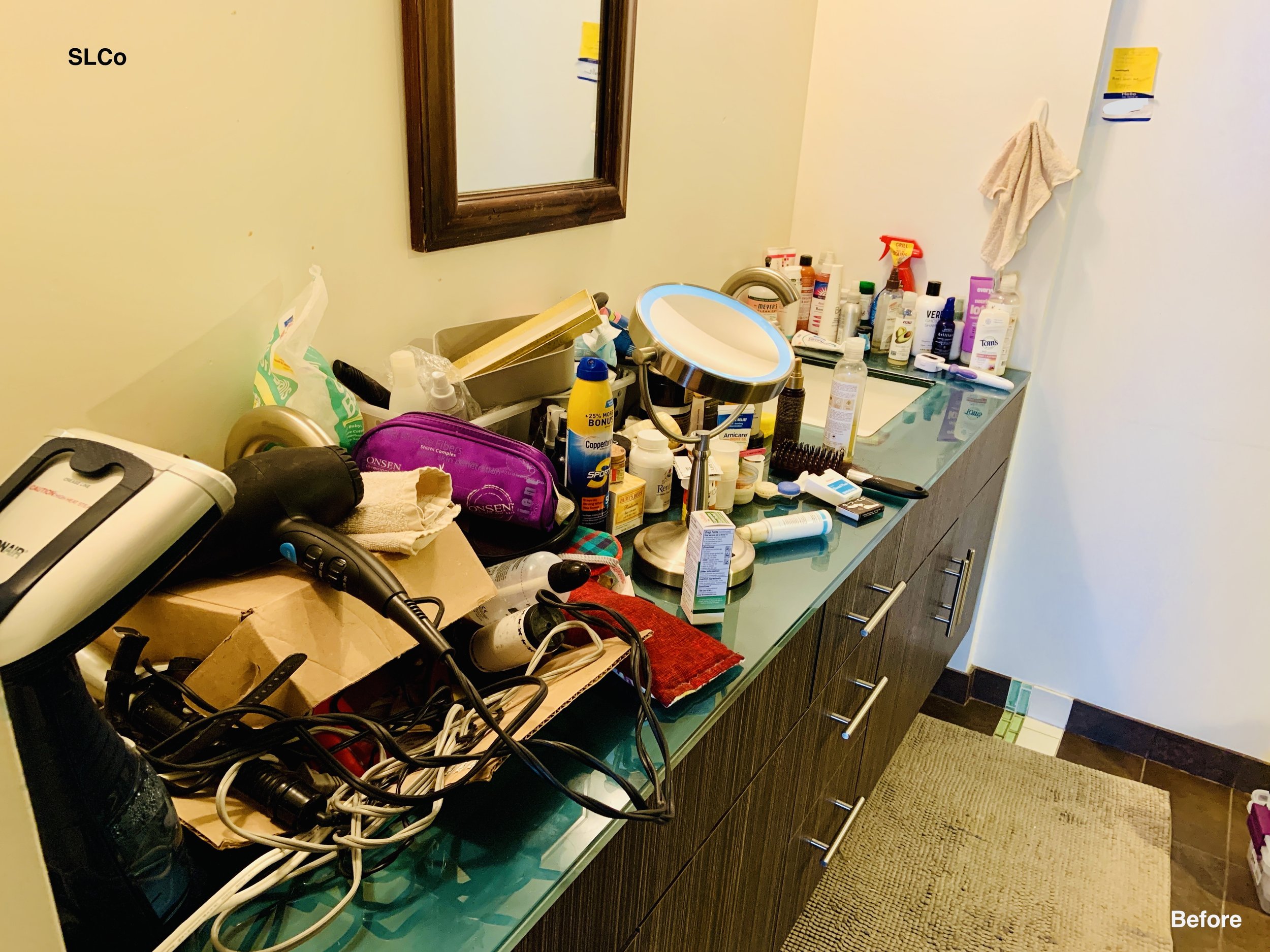 Before photo of a bathroom with boxes, appliances and items stacked on bathroom counter and in one of the two sinks, unusable space.