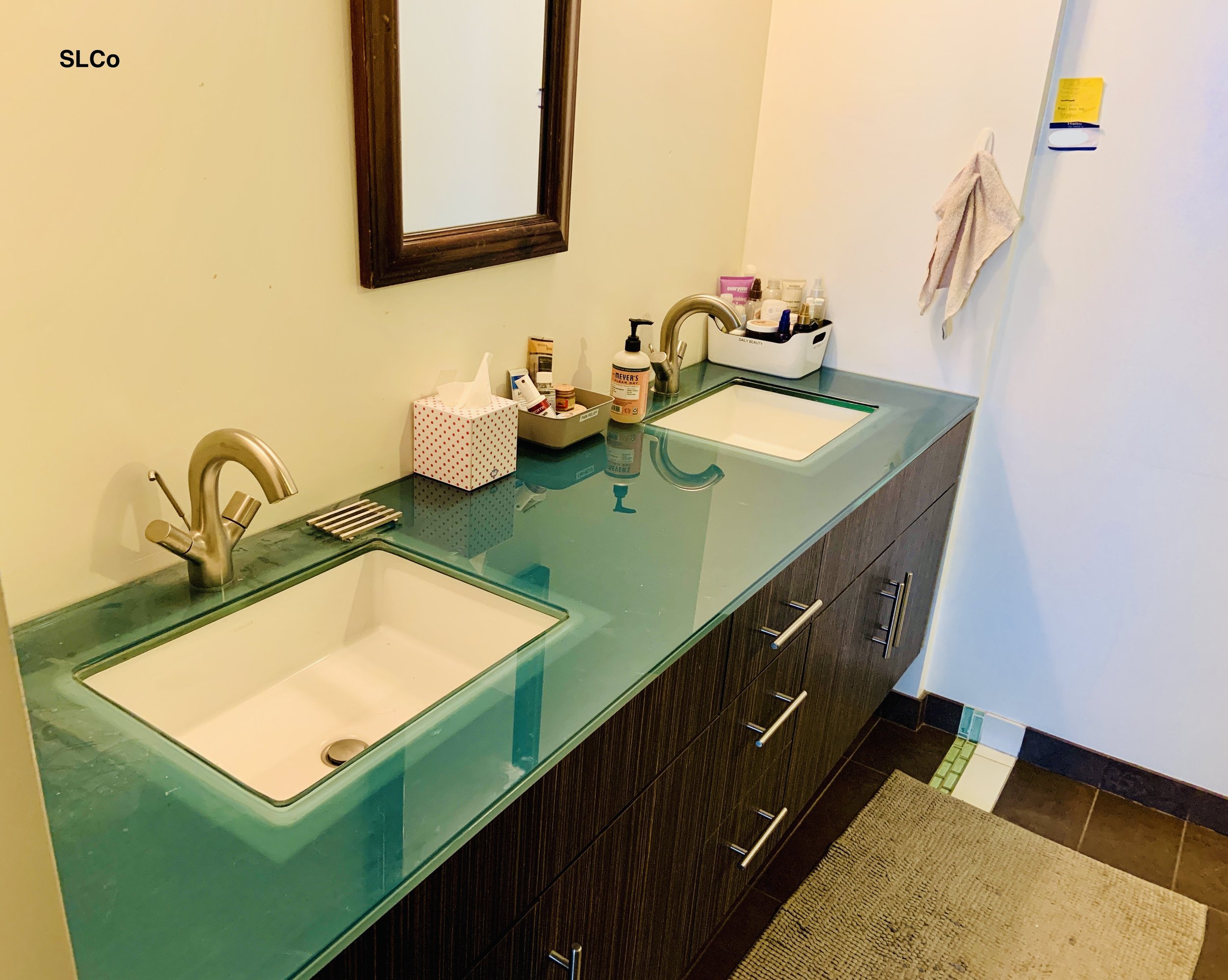 After photo of bathroom counter with very few items on counter, can see trans-blue glass counter on dark wooden shelves, and see both sinks.