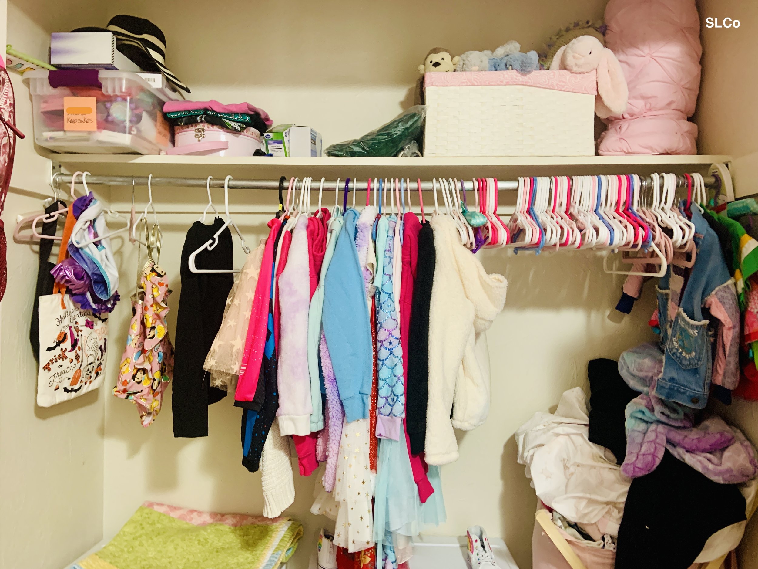 Little girls closet with clothes hanging in center of closet and shelf on top with items falling on each other