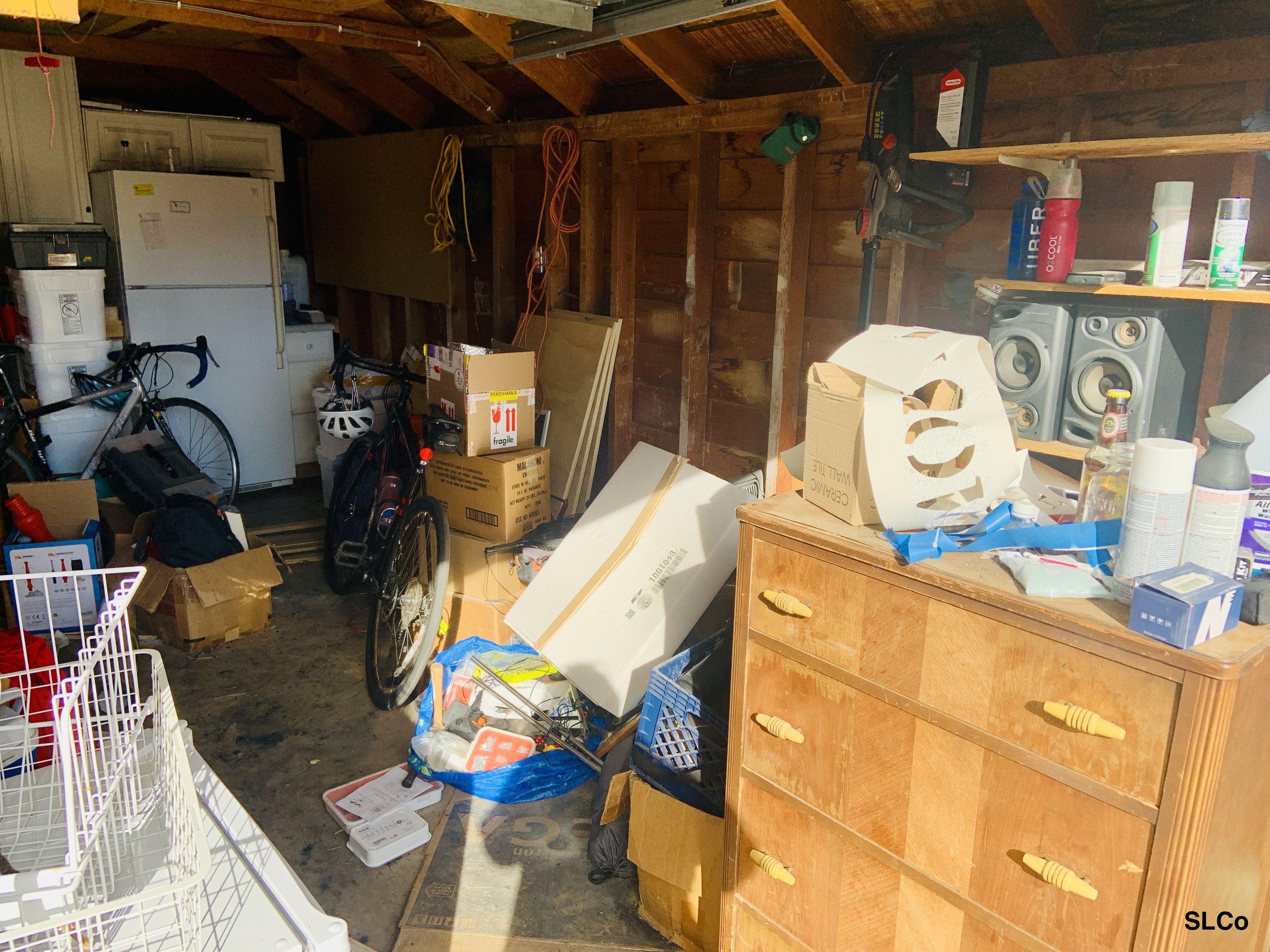 Before photo of garage with mattress leaning on top of items, fridge in middle of floor, and dresser in middle, boxes on floor