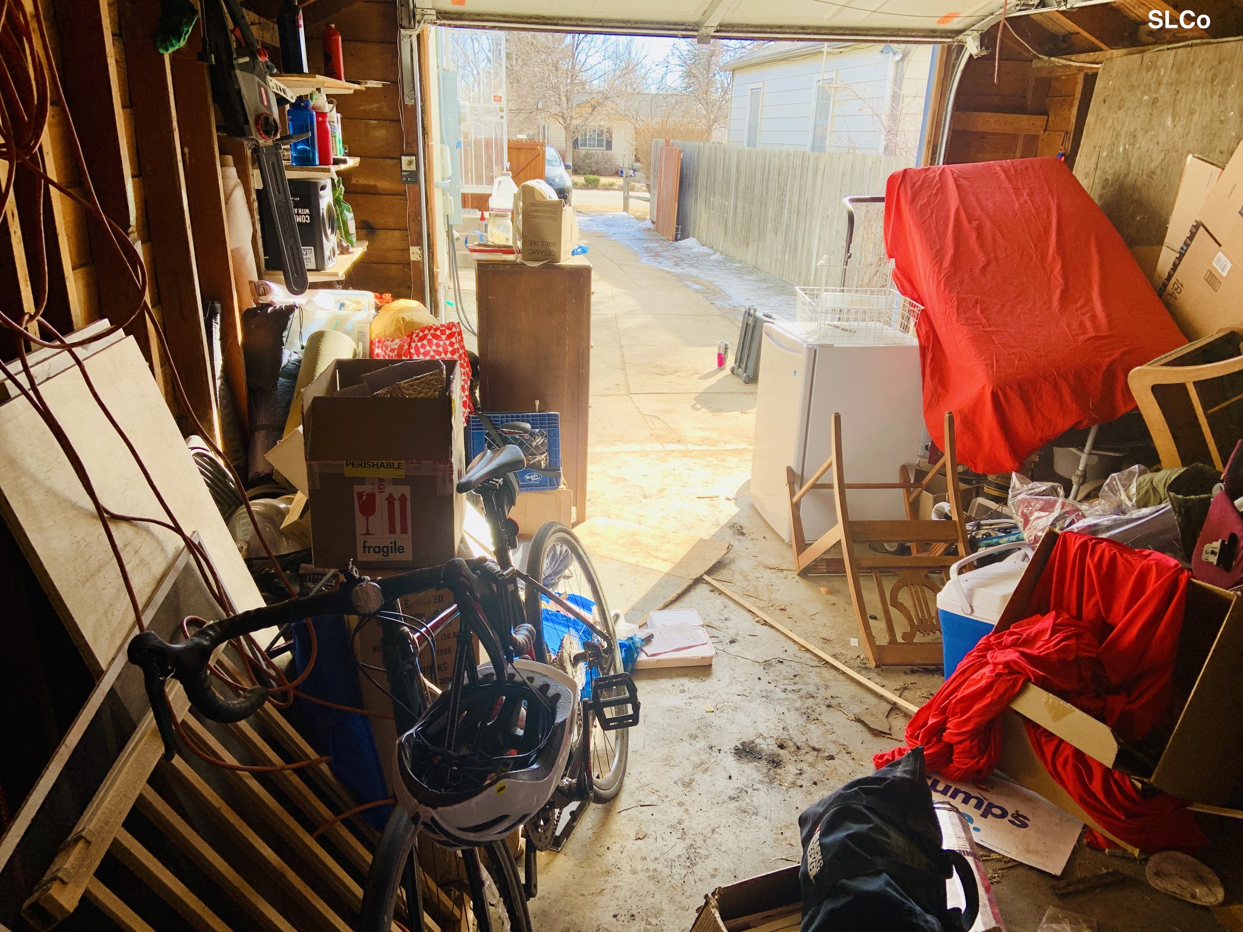 Before photo of garage with mattress leaning on top of items, fridge in middle of floor, and dresser in middle, boxes on floor