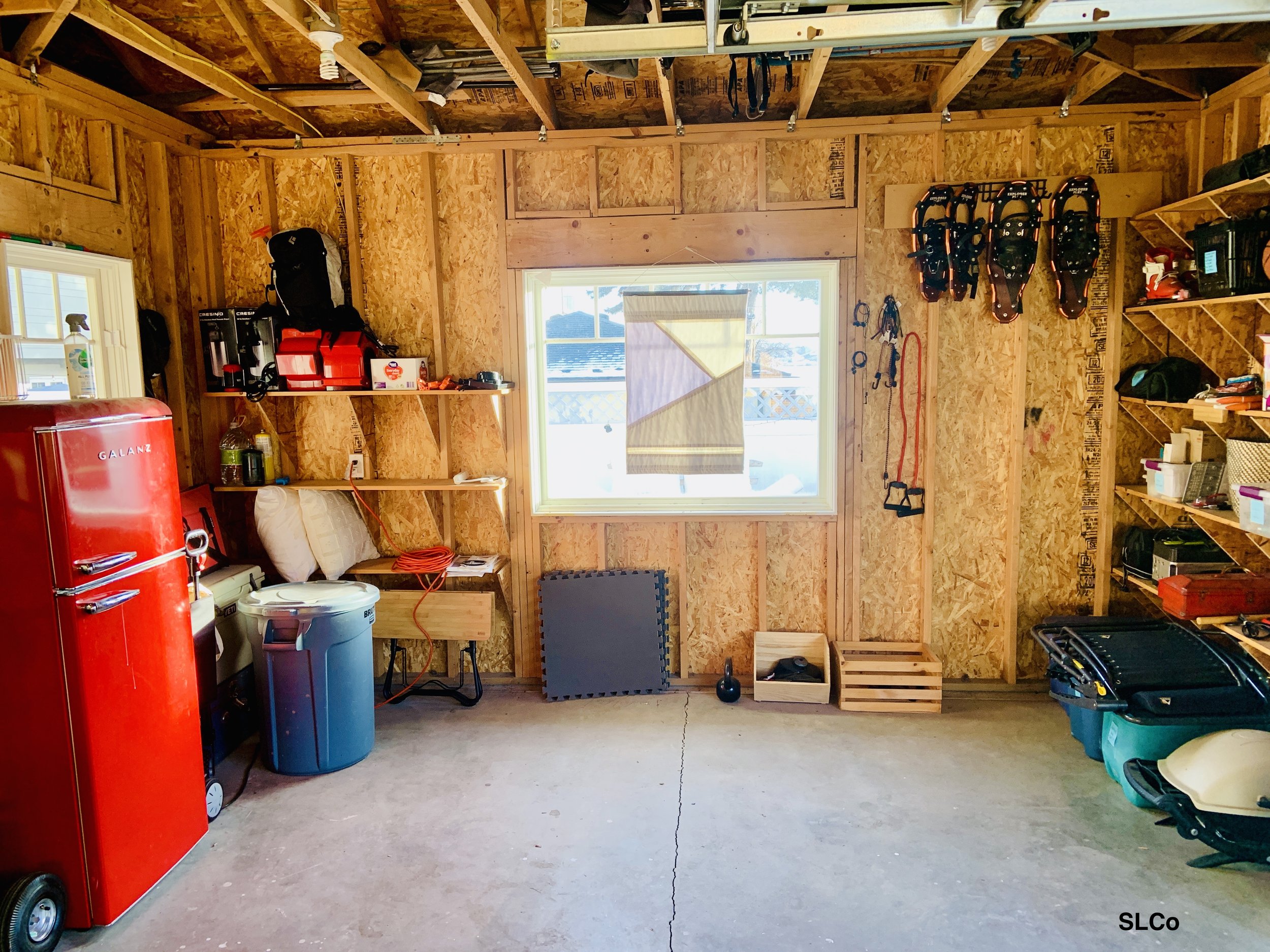 Before photo of garage with wooden shelves on the side, red fridge on the side and floor clear