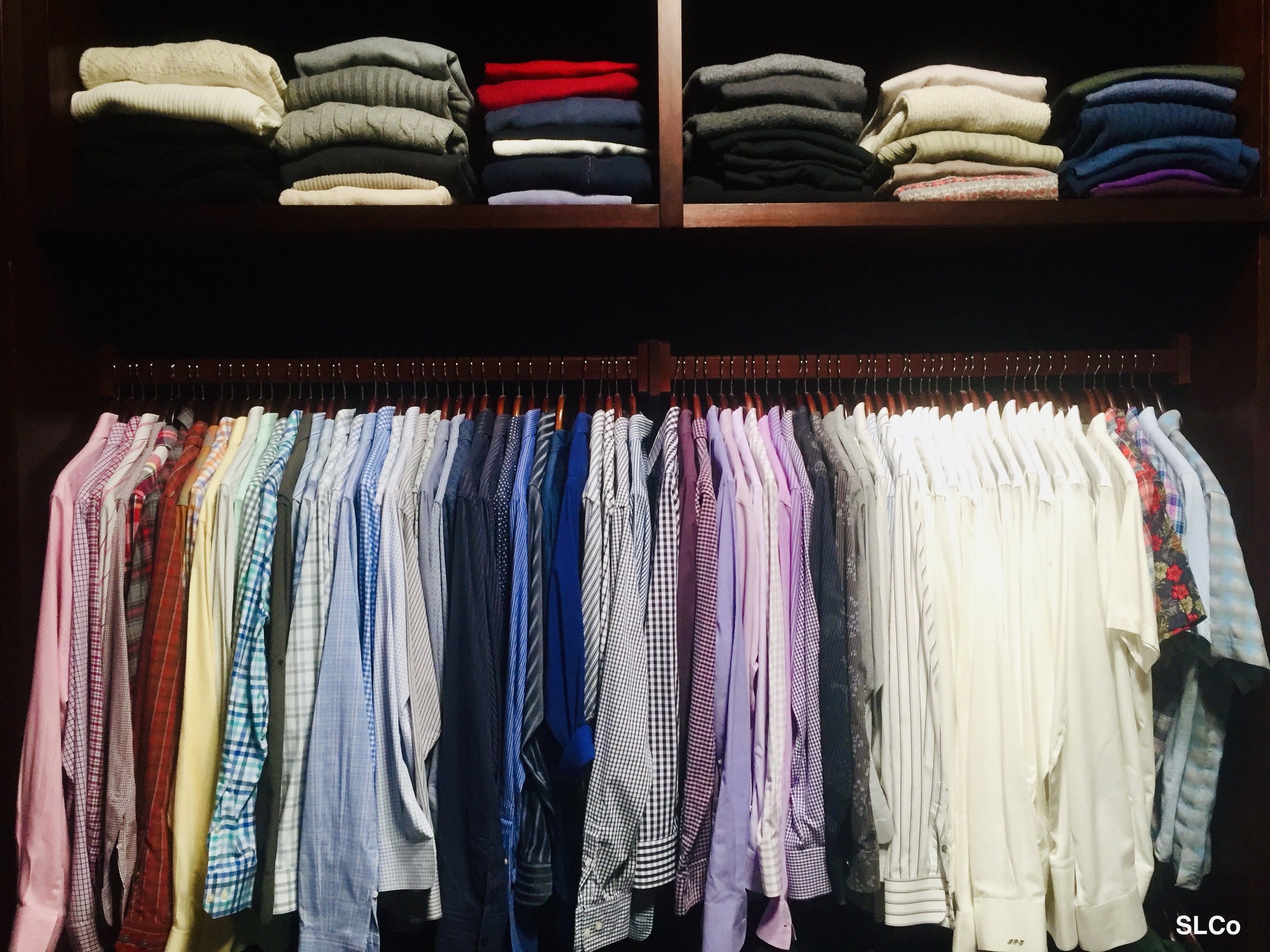 Large mens closet organized with shirts hanging with all same colors together on two rows of hanging dress shirts on bars