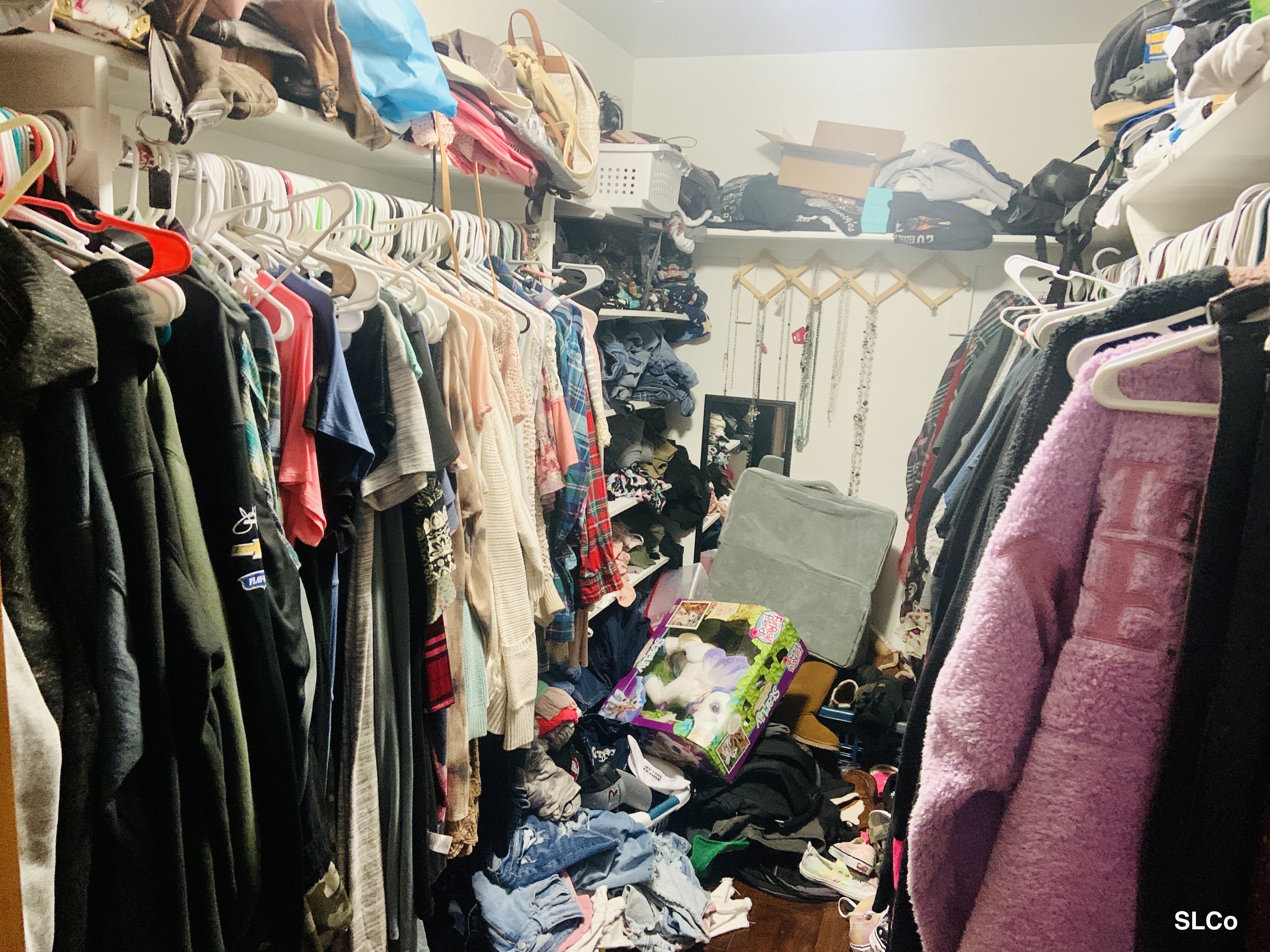 Cluttered closet with clothes hanging and packed in and clothes and items all over floor of walk-in closet.