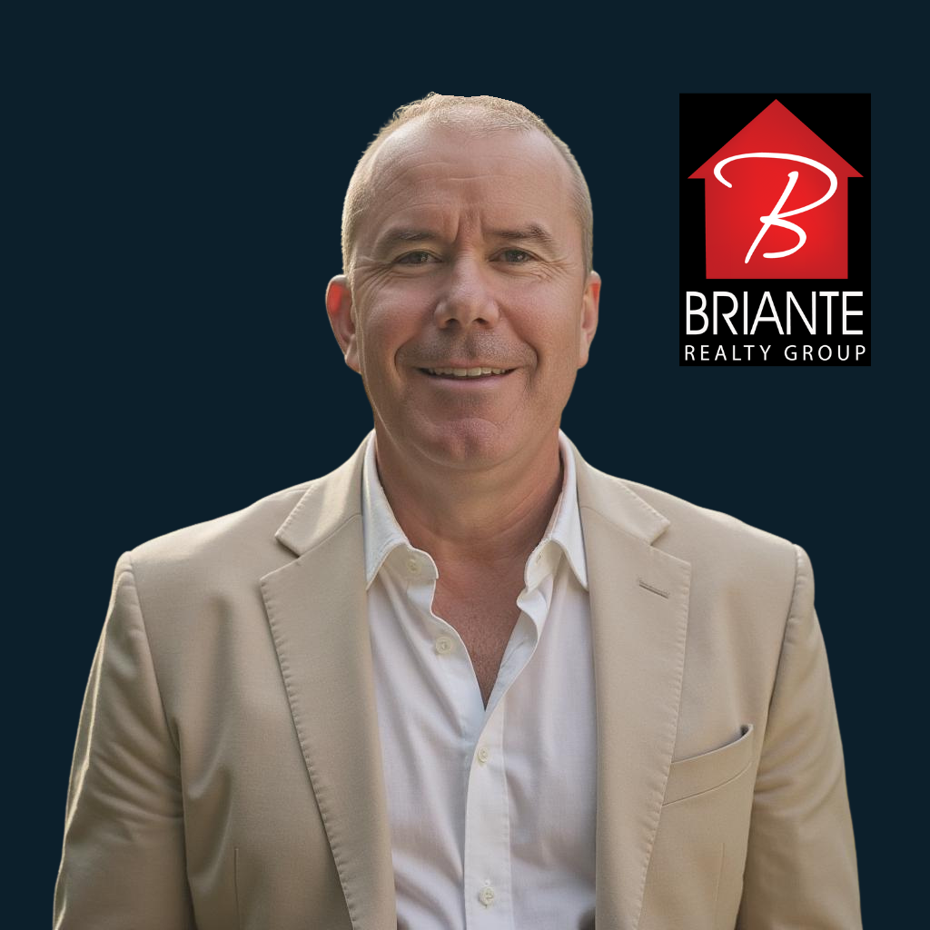 A man in a beige suit jacket and white shirt smiling in front of a dark background with a real estate logo.