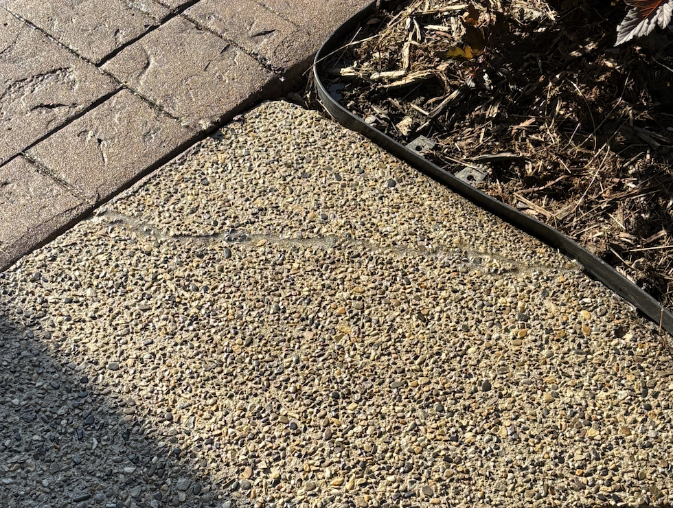 Close-up of a textured concrete sidewalk with a rock and pebble surface, adjacent to a paved brick pathway and a garden bed with mulch.