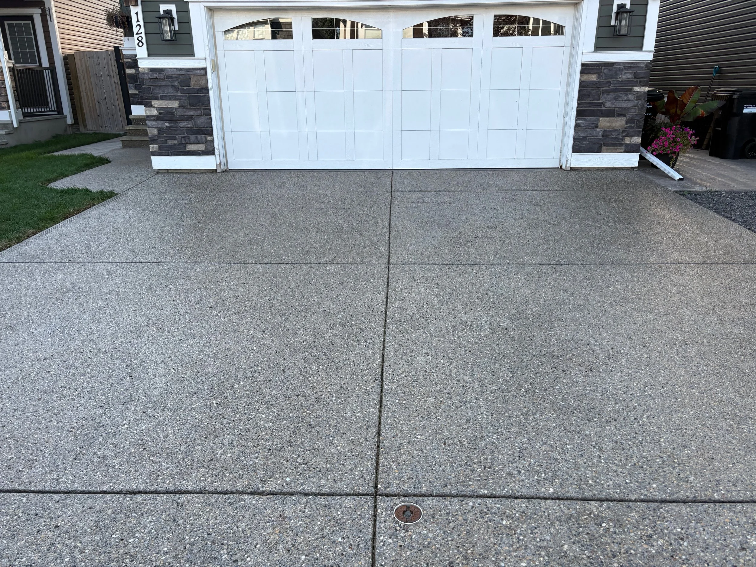 Concrete driveway with a drain cover in front of a white garage door. To the left is a small staircase with a stone base leading to a side door, and to the right is a small garden with potted plants and trash bins.