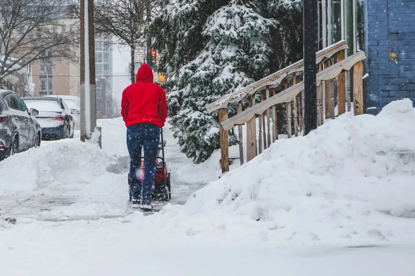 Person in a red hoodie shoveling snow on a city sidewalk during a snowstorm.