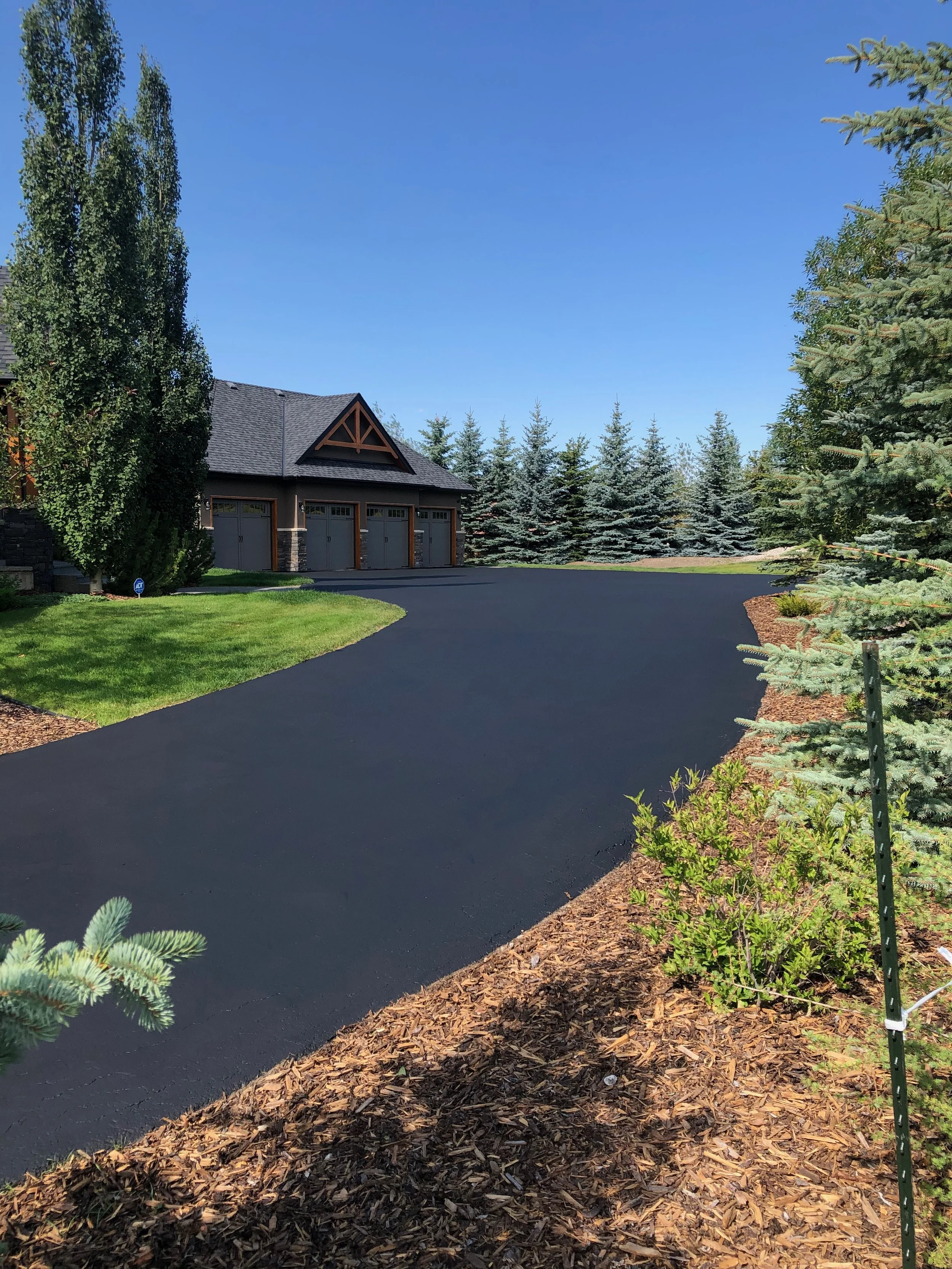 Freshly paved black asphalt driveway leading to a house with three garage doors, surrounded by green grass, trees, and shrubs under a clear blue sky.