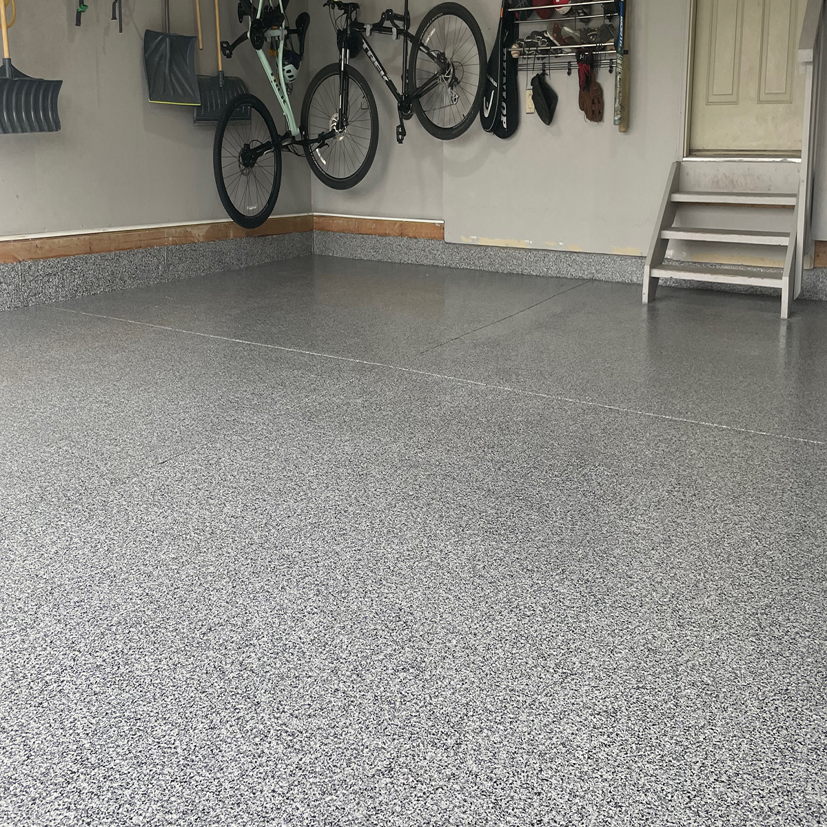 Empty garage with textured gray floor, wall-mounted bicycle rack holding two bicycles, and storage hooks with tools and items on the wall.