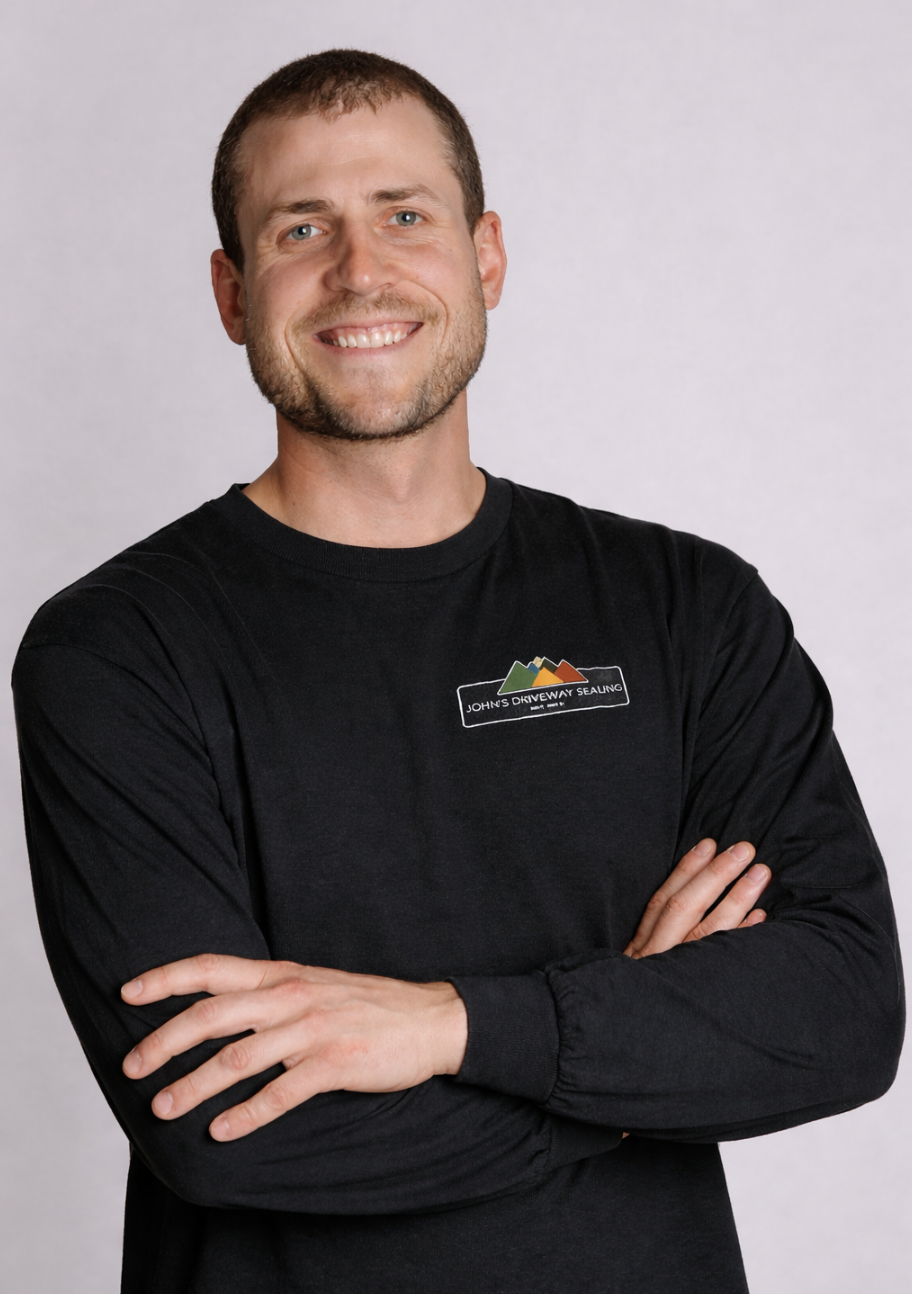A smiling man with short brown hair and a beard, wearing a black long-sleeve shirt with a logo that says 'John's Driveway Sealing' and has a mountain graphic, stands with crossed arms against a plain light gray background.