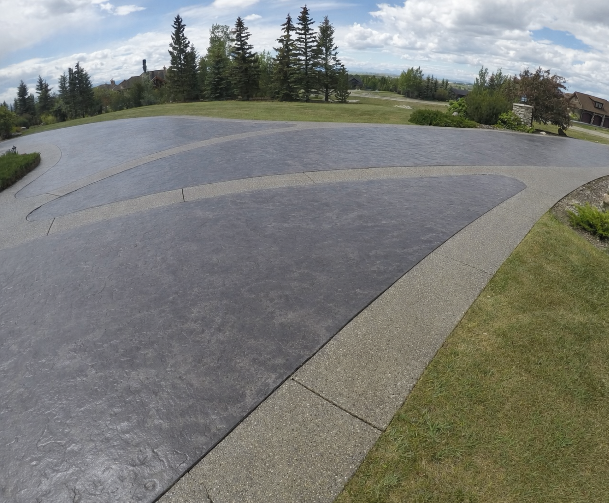 A residential driveway with dark-colored and light-colored concrete pavers, surrounded by a lawn and landscape, with trees and houses in the background under a partly cloudy sky.