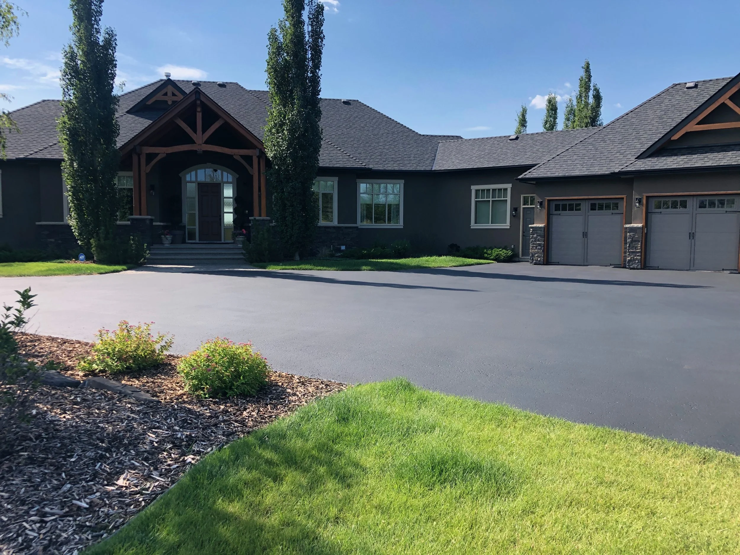A modern house with a large driveway, green lawn, and landscaped garden, featuring a dark gray exterior and multiple garage doors.