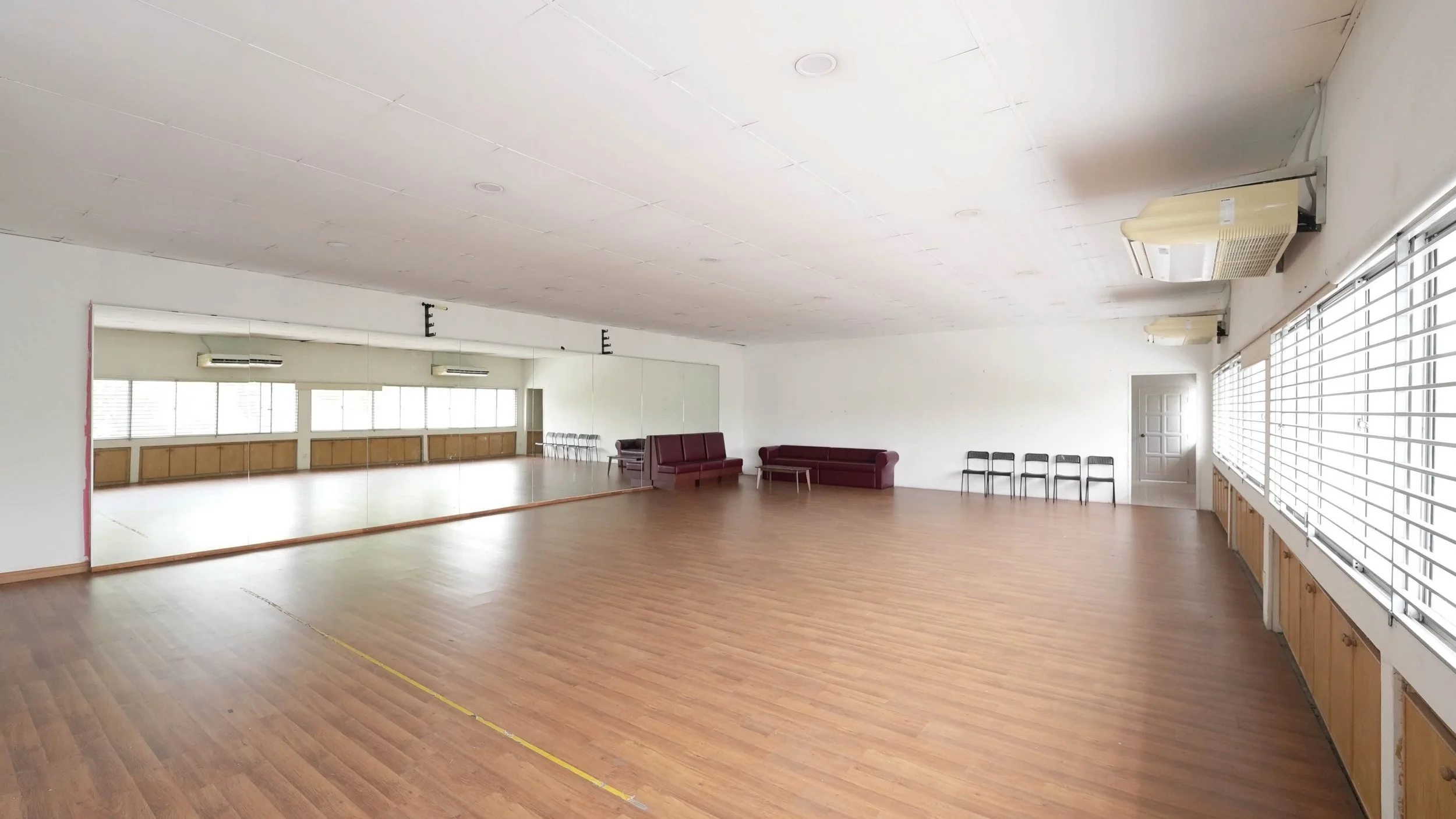 Empty ballroom or dance studio with large mirror wall, wooden floor, rows of chairs, and windows with blinds.