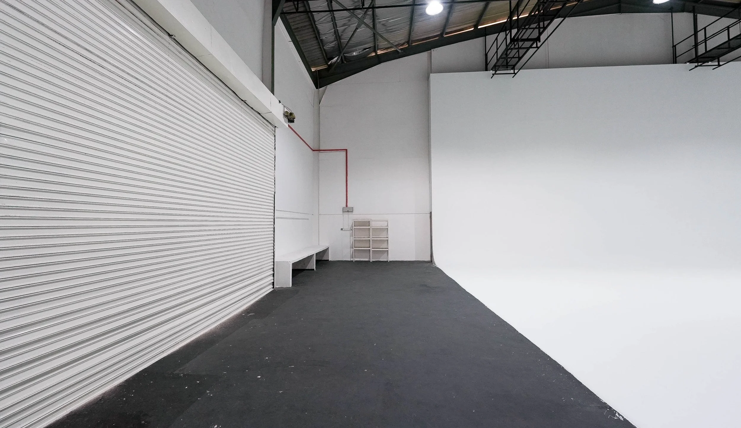 Empty industrial space with a closed white rolling shutter door on the left, white walls, a black floor, a metal staircase above, and a small white shelving unit at the back.