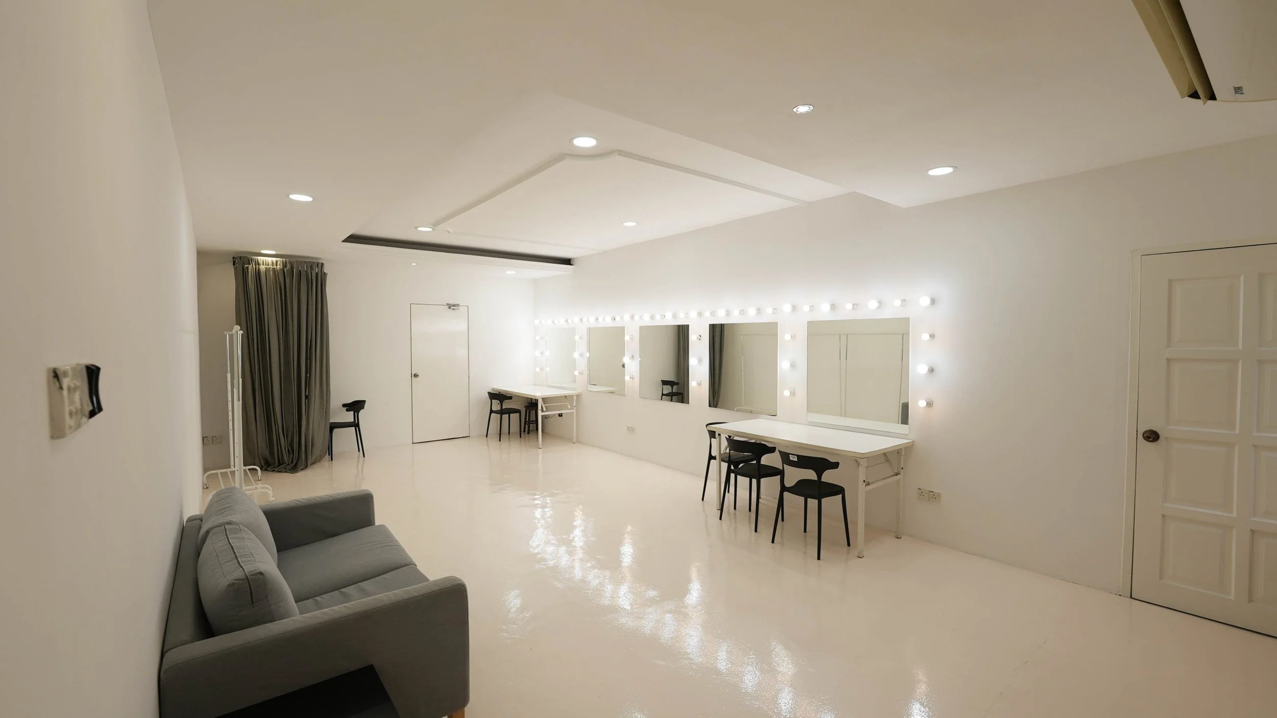 A brightly lit room with white walls, three mirrors with lights around them, black chairs, a gray sofa, and a white table, arranged for makeup or dressing.
