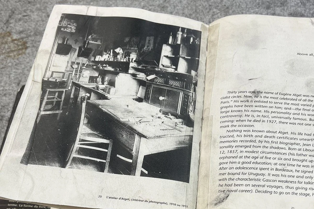 An open book with a photo of the photographer's studio ahowing a desk, chair and stool in a cramped room with bottles and many objects on the shelves near the desk