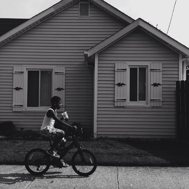 A young boy riding a bicycle on a sidewalk in front of a house with two windows and shutters, holding a drink.
