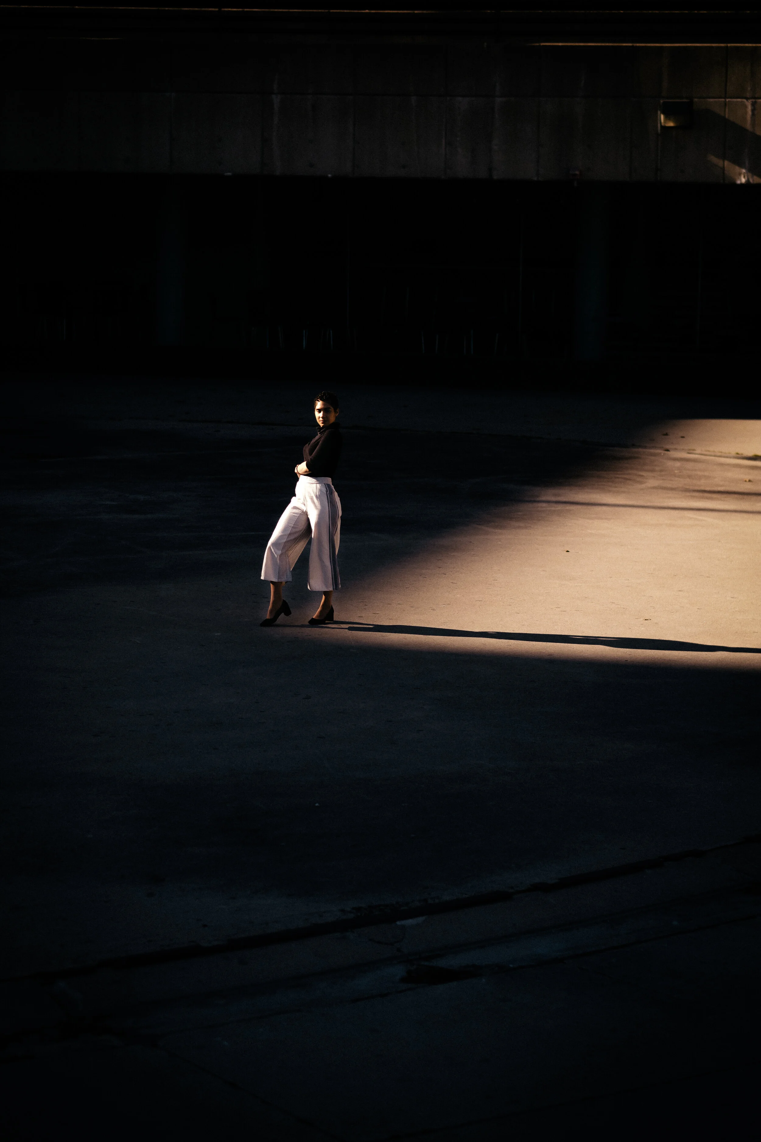 A woman stands alone on a dark street illuminated by a beam of light, with a shadow cast behind her.