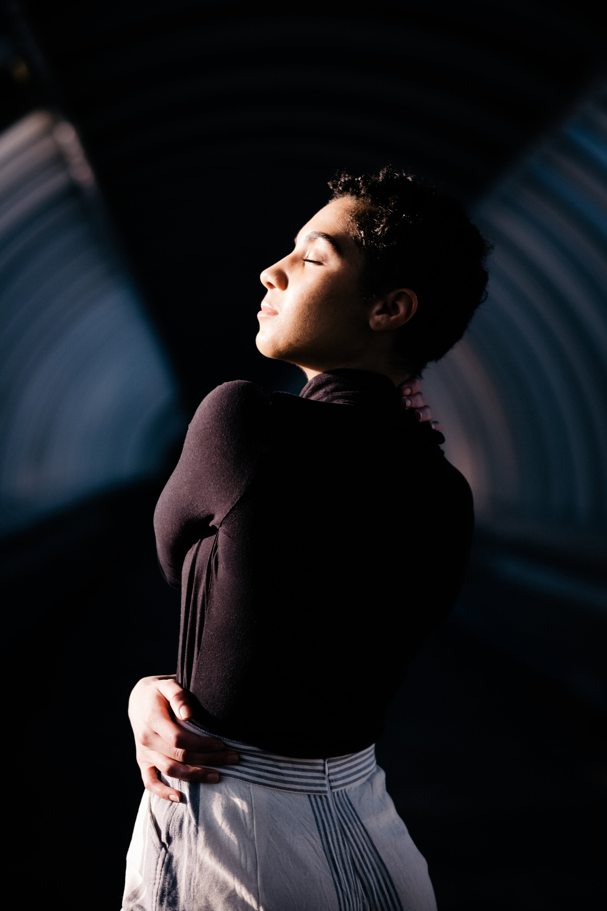 Young person with eyes closed, standing with arms crossed, in a dark tunnel with blue lighting.
