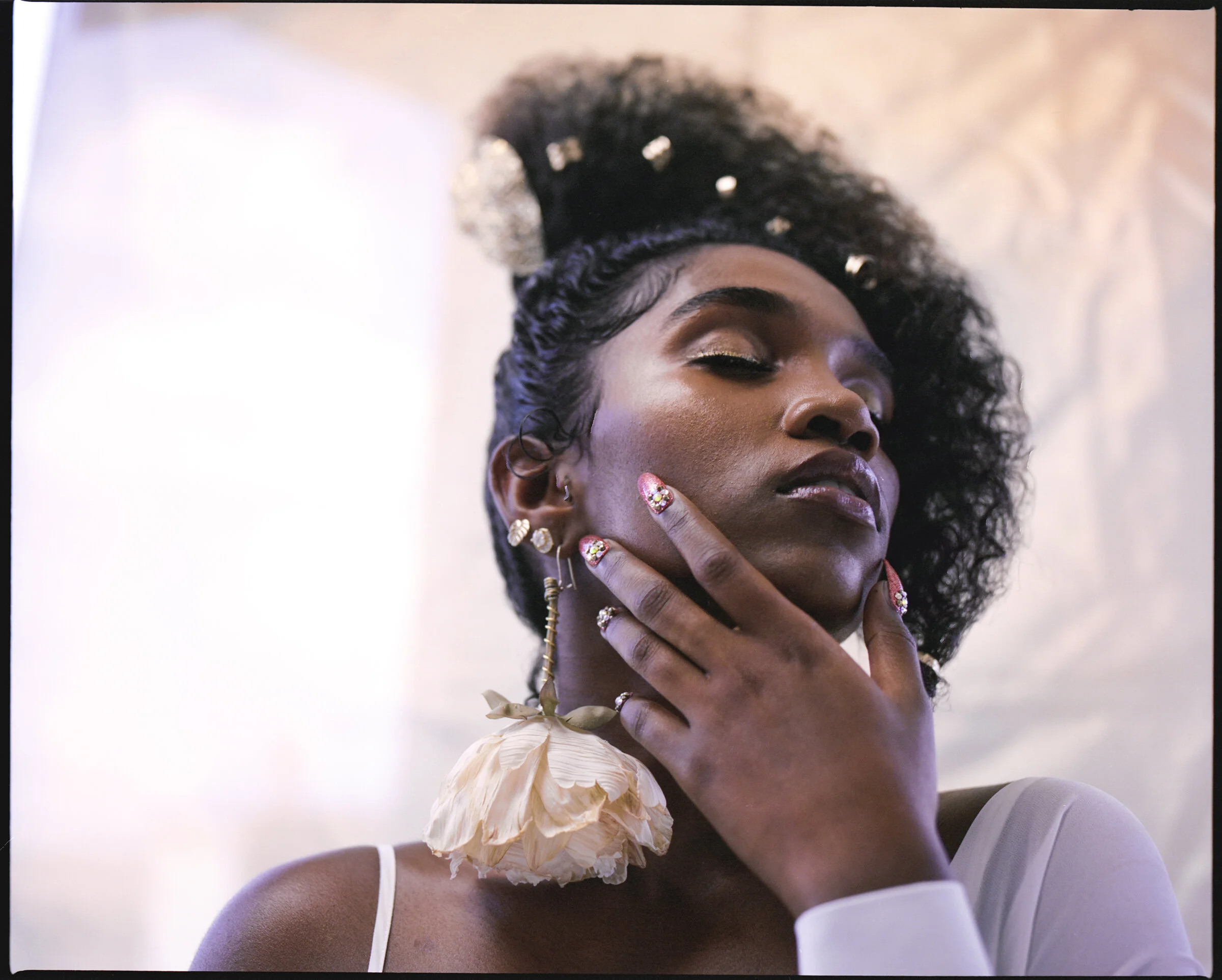 A woman with dark curly hair adorned with pearls, wearing gold eyeshadow and jewelry, and a large decorative flower earring. She is touching her face with her hand, confidently posing against a neutral background.