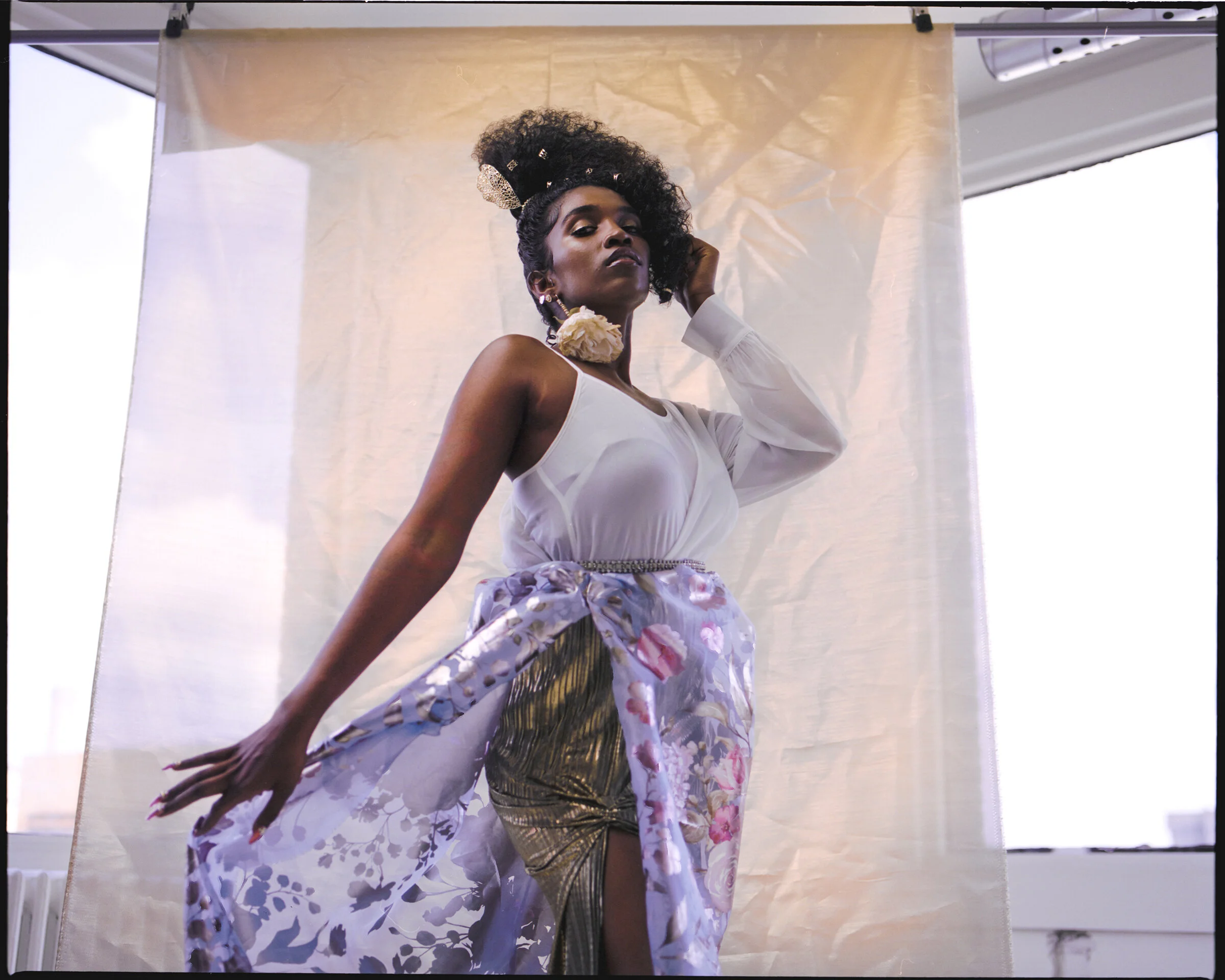 A woman with styled hair and accessories posing in a white top, a floral robe, and metallic gold pants in front of a beige backdrop.