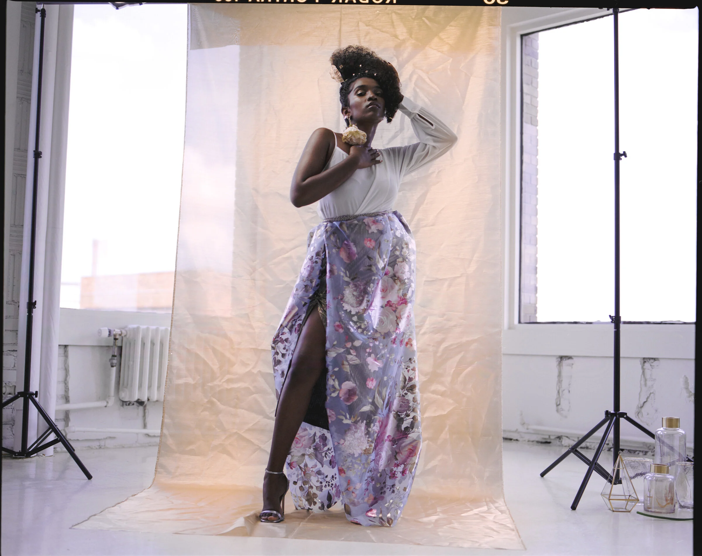 A woman with dark skin and curly hair posing in a studio with natural light, wearing a white top, a long floral skirt with a high slit, high heels, and jewelry, holding a flower near her neck, in front of a beige backdrop, with studio equipment and w