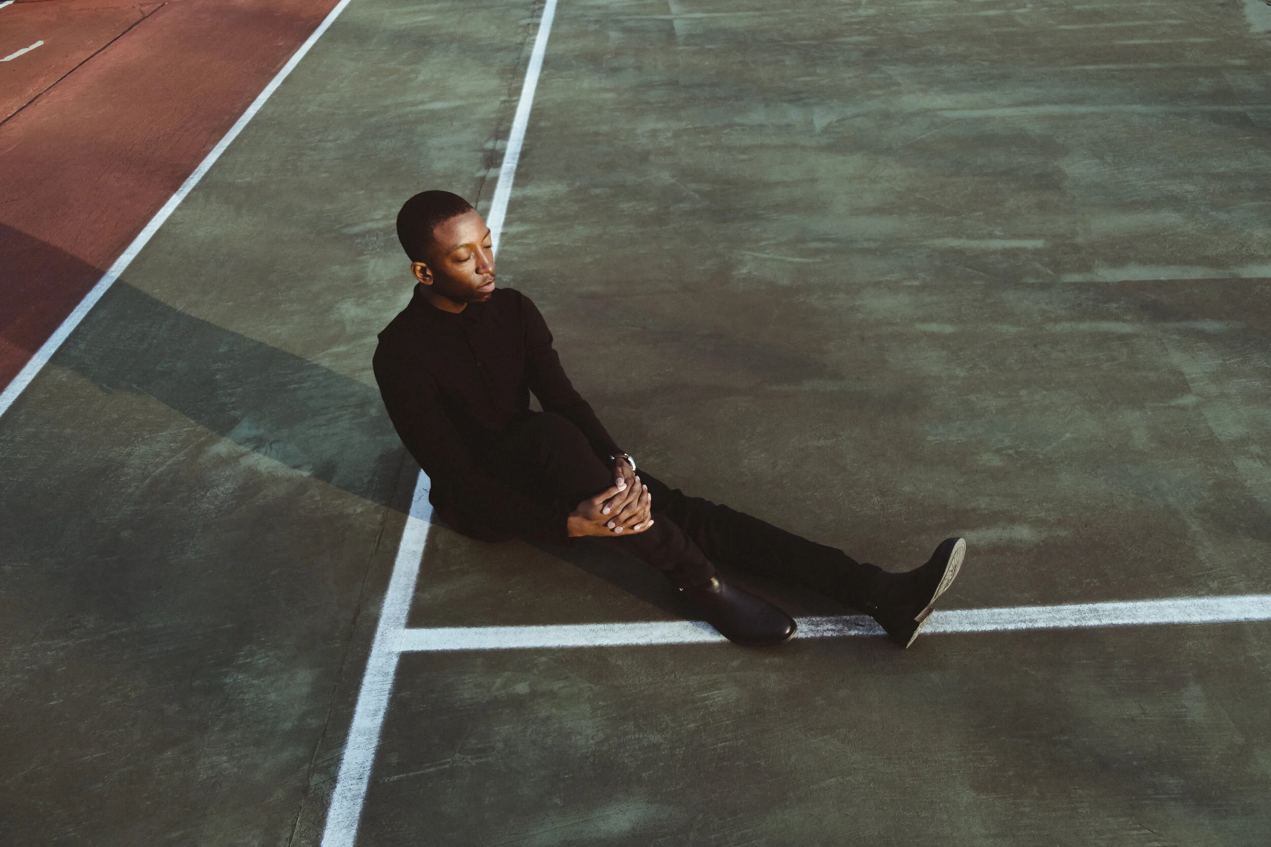 Person sitting on a sports court with eyes closed, dressed in black, leaning against a corner and appearing contemplative.