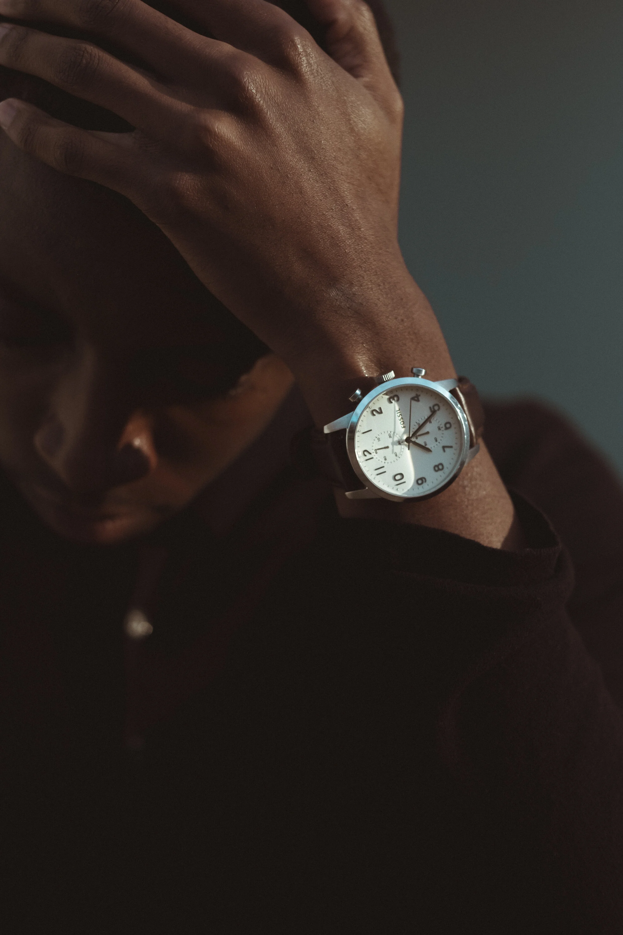 A person wearing a wristwatch with a white face and brown strap, resting their hand on their face, with a dark background.