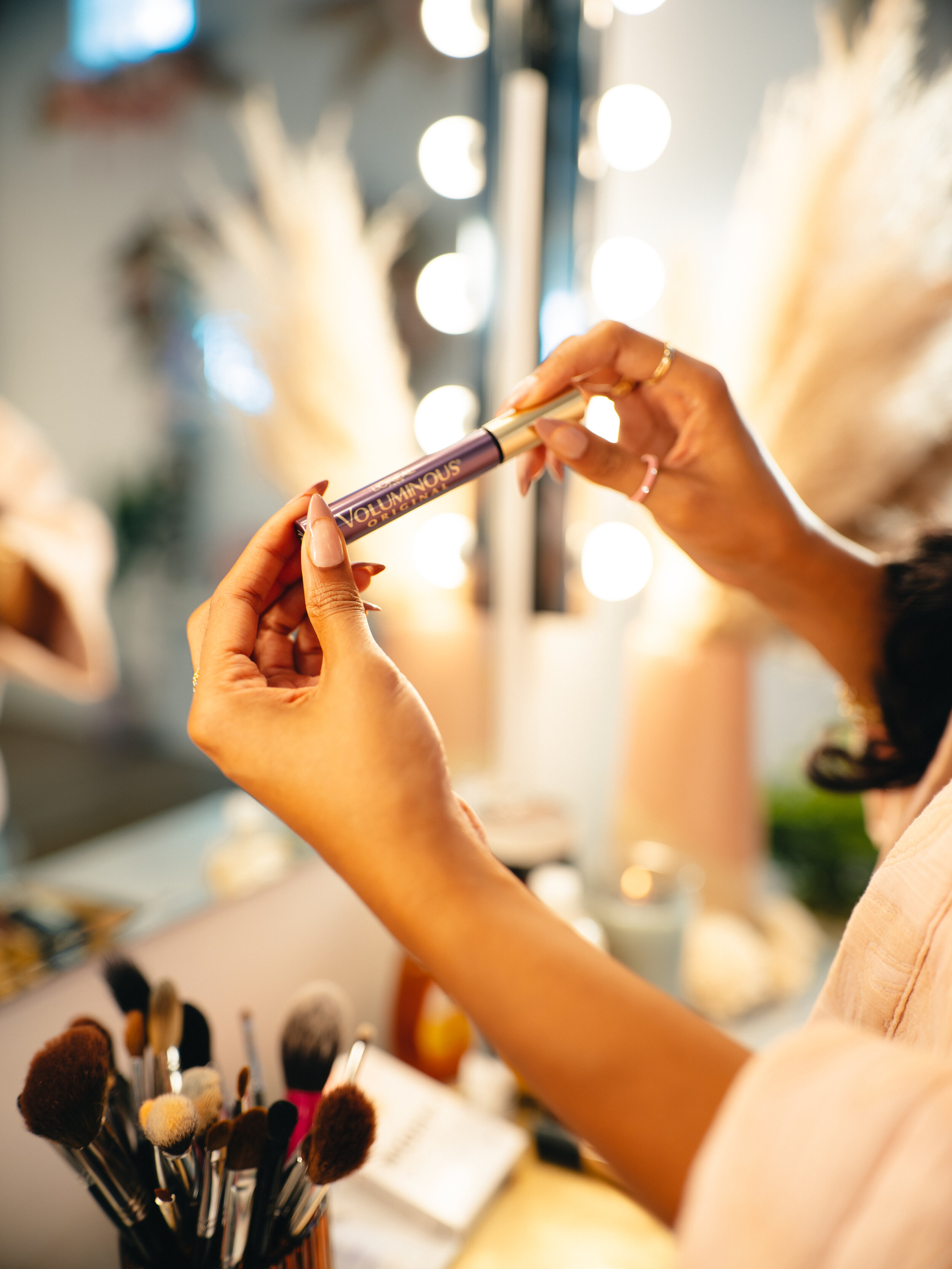 A person holding a tube of volumizing mascara in a makeup studio with bright lights and makeup brushes in the foreground.