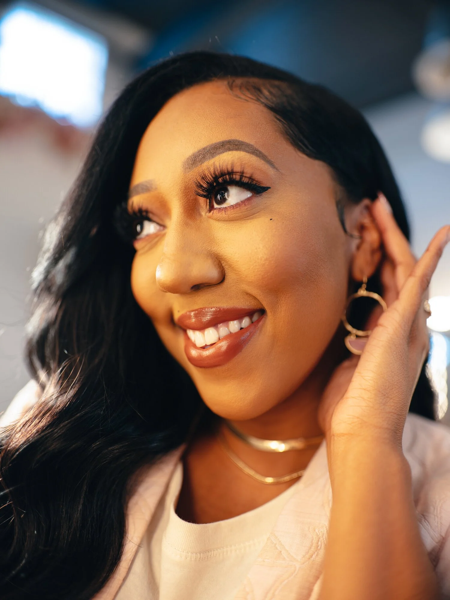 A young woman with long black hair, makeup, and earrings smiling and touching her ear.