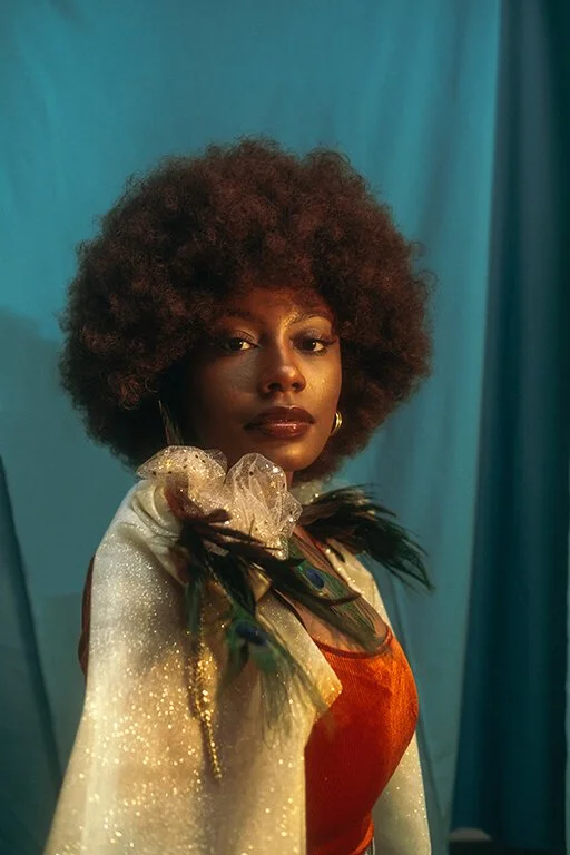 A woman with a voluminous curly hairstyle and gold hoop earrings, dressed in a shiny gold jacket with feather accents on the shoulders, and an orange top, posing in front of a teal curtain.