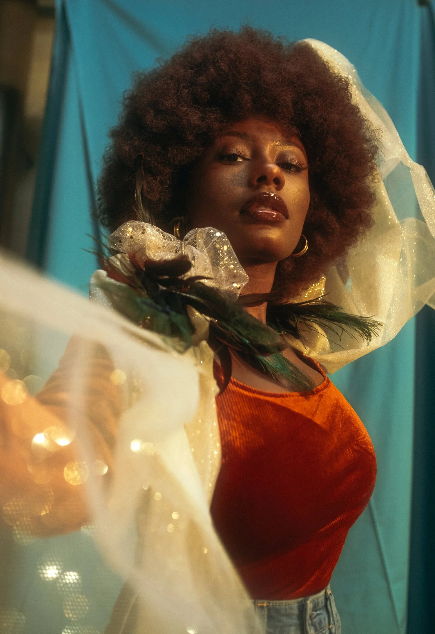 A woman with curly hair and dark lipstick posing with a blue background, wearing a red top, accessorized with feathers and glittery fabric in a fashion shoot.