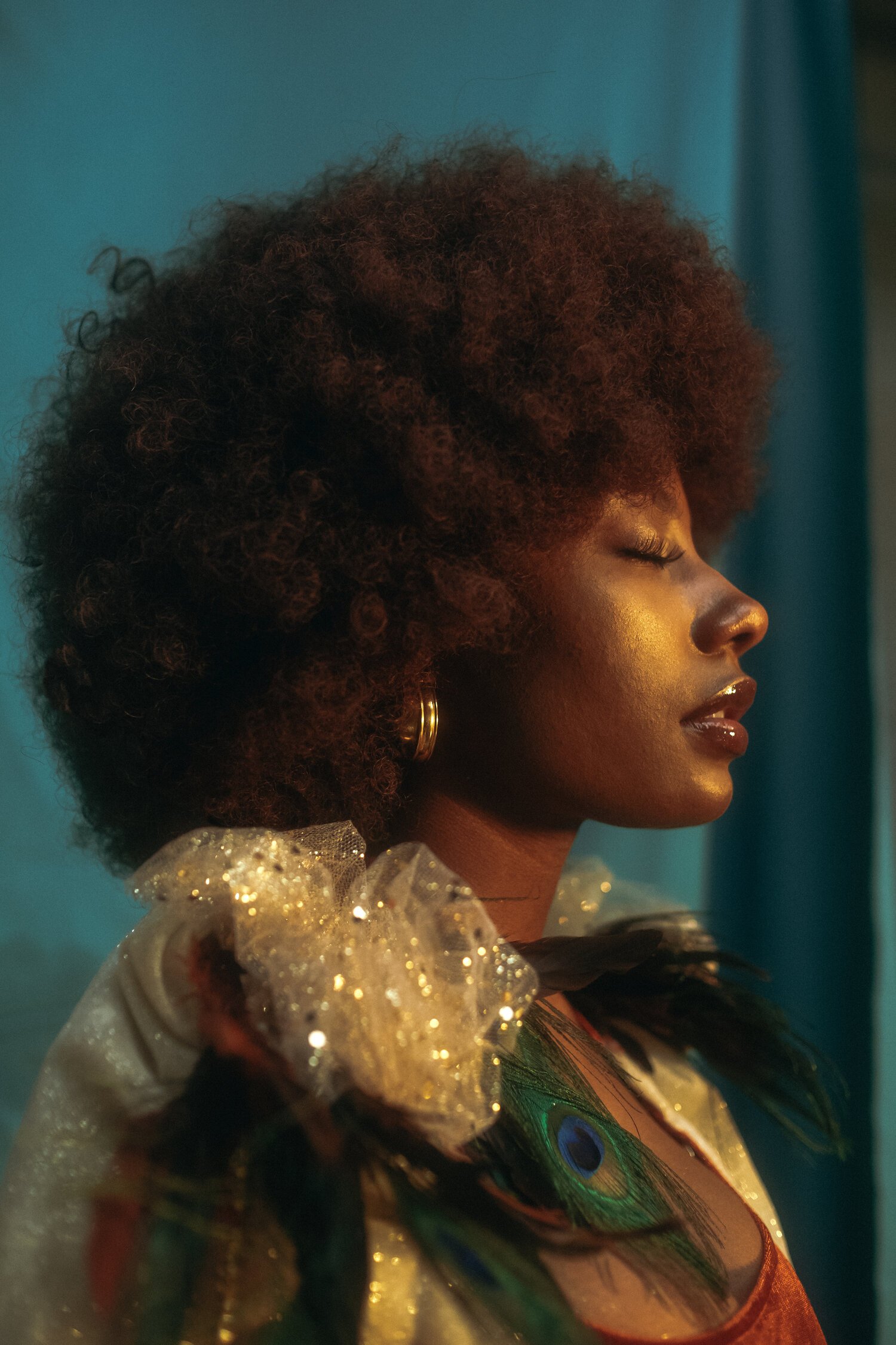 A woman with curly, voluminous hair, wearing gold earrings, a shimmering top with glitter, and peacock feather decorations on her outfit, standing with eyes closed against a blue background.