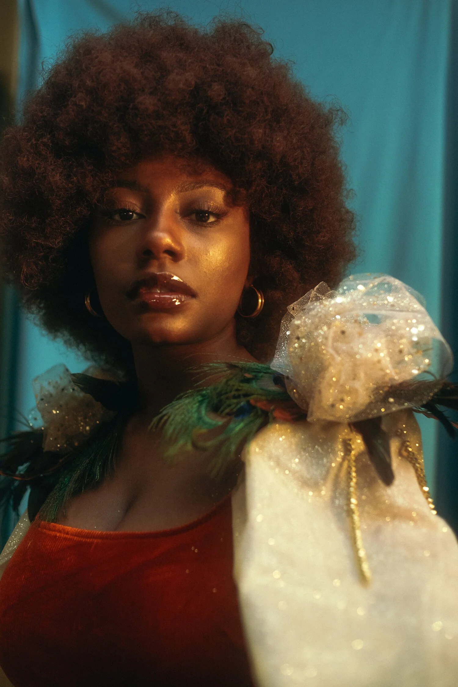 Close-up portrait of a woman with dark skin, curly hair, gold makeup, and shiny lips, wearing a glamorous dress with feather and glitter embellishments, and gold hoop earrings, against a blue background.
