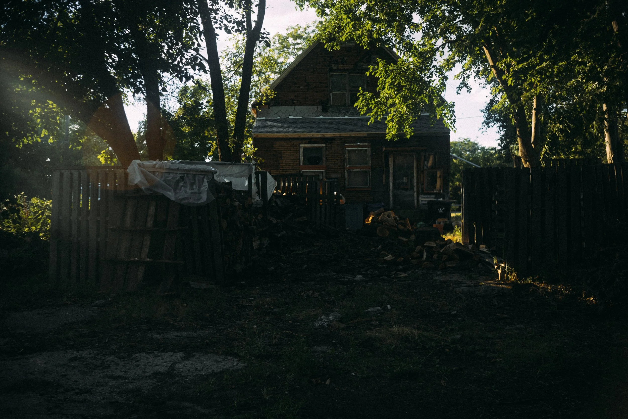 A dark, abandoned house with broken windows and a damaged roof, surrounded by a worn wooden fence and trees, with a pile of wood and debris in the yard.