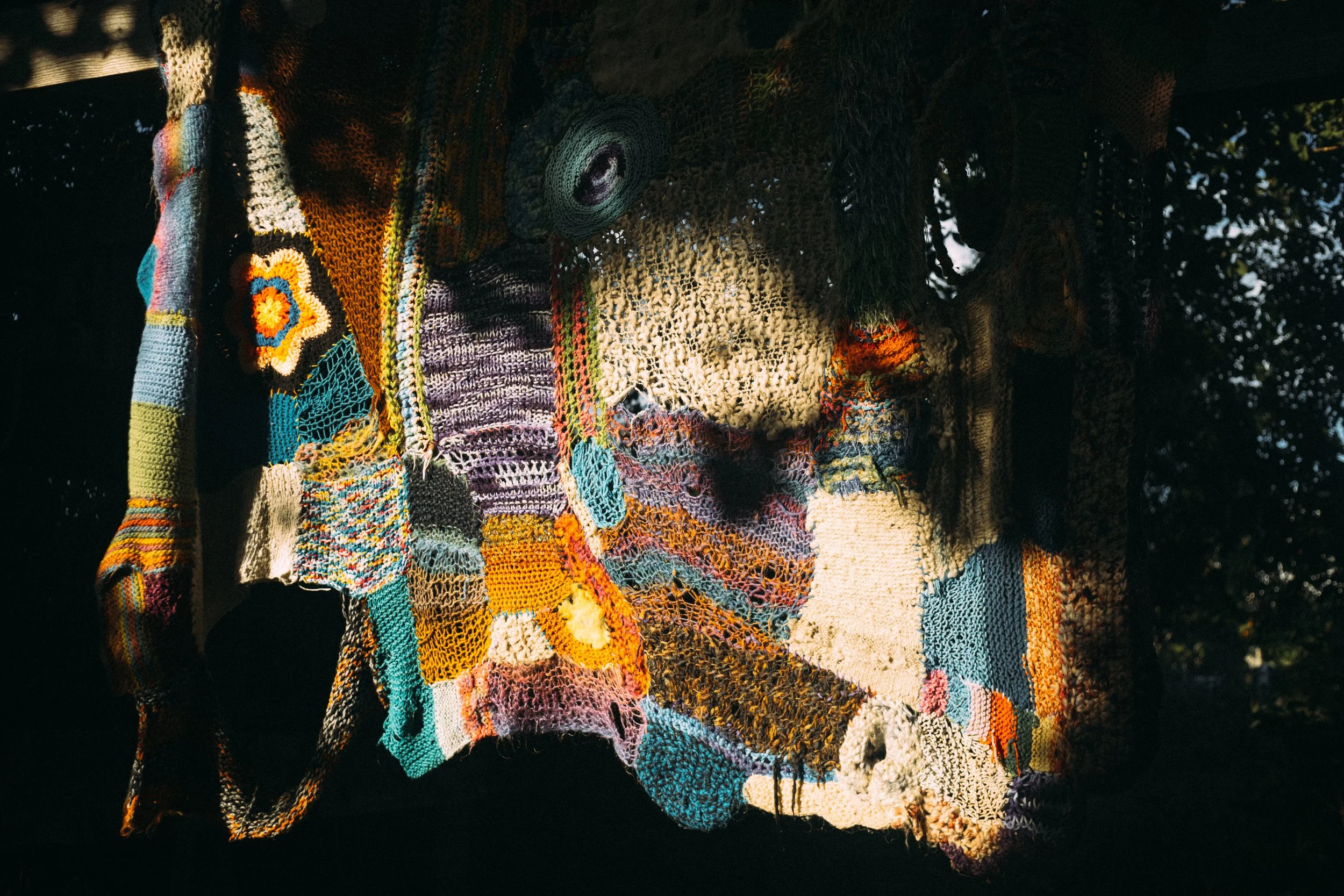 Colorful patchwork textile hanging outdoors with sunlight filtering through trees.