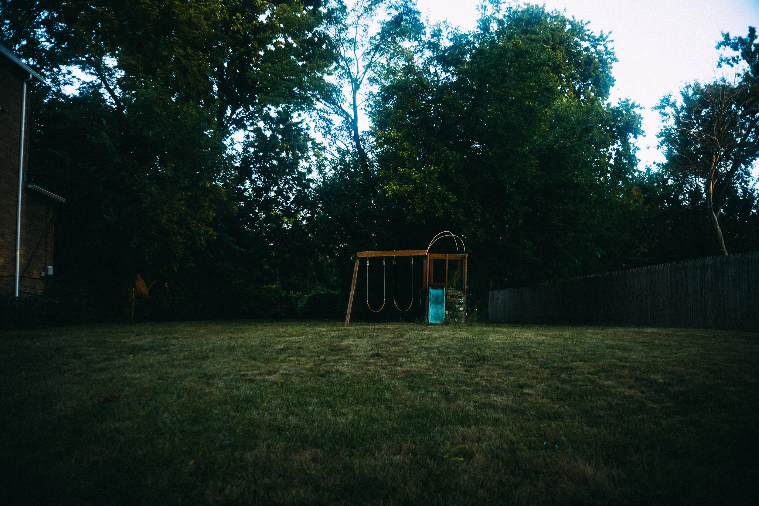 Empty backyard with a wooden swing set, a slide, and a fenced yard with large trees in the background during dusk.