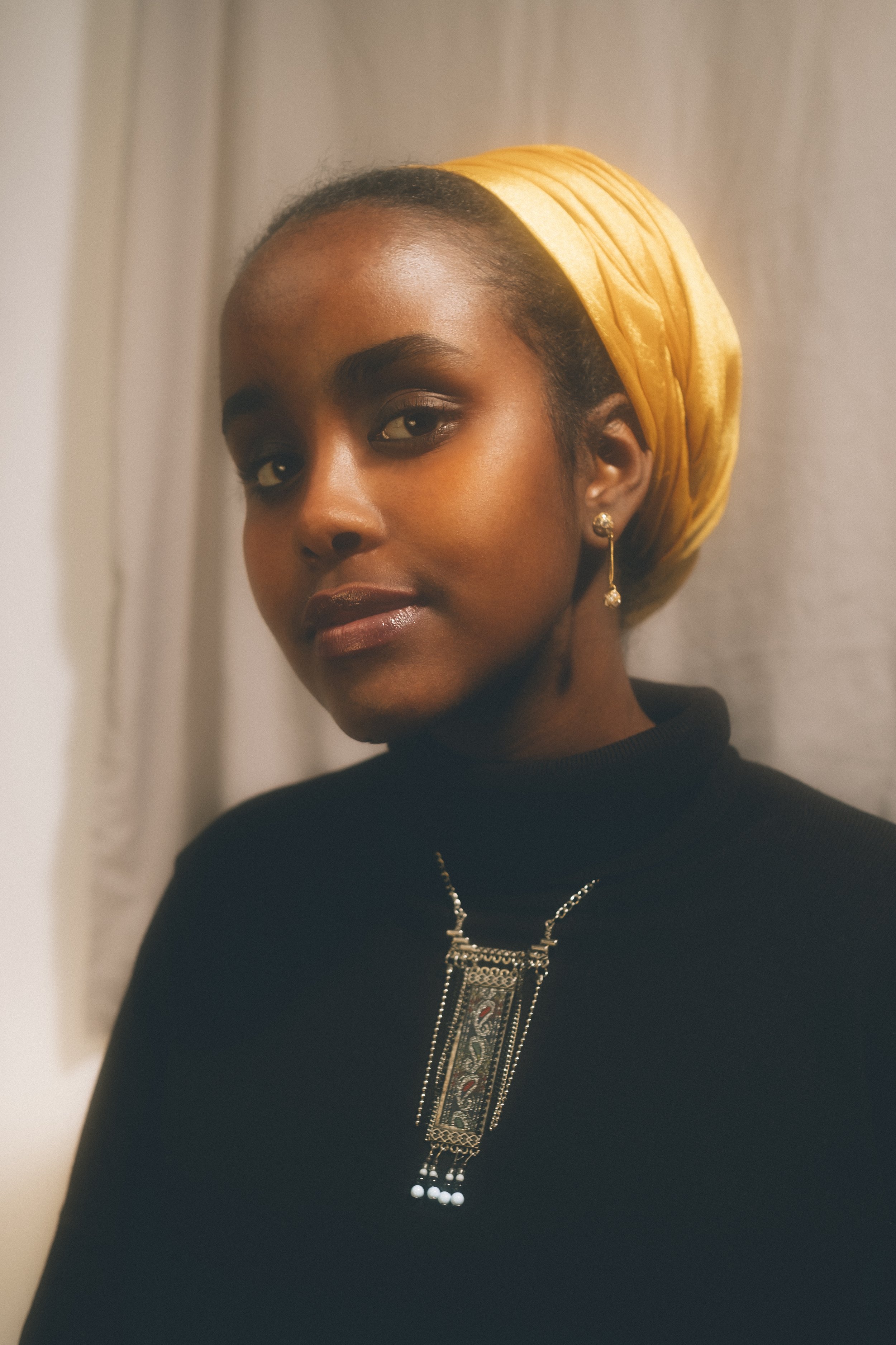Portrait of a woman wearing a yellow headscarf, black top, and jewelry, standing in front of a light-colored curtain.