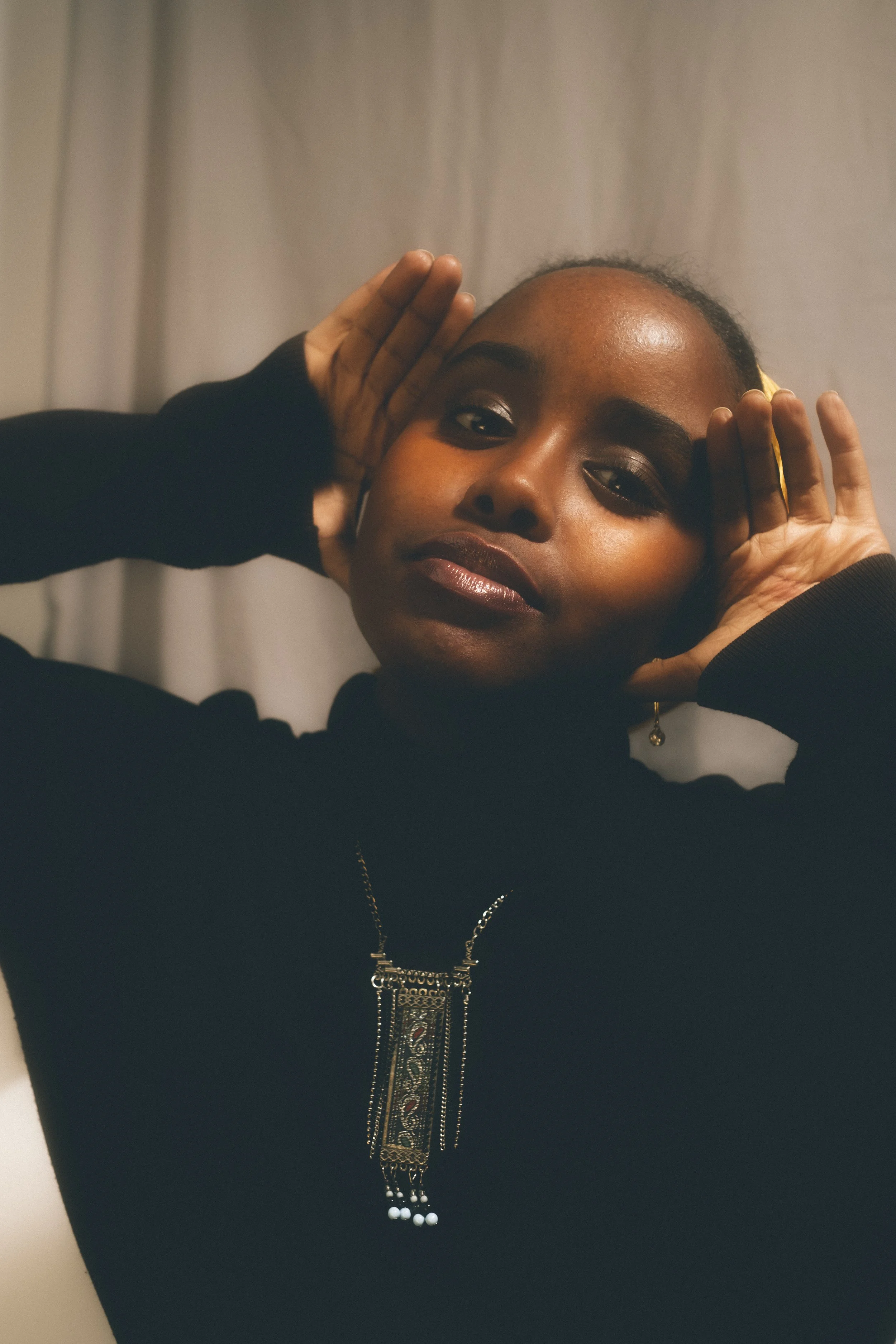 A woman with short hair wearing a black top, gold jewelry, and touching her head with both hands, looking at the camera.